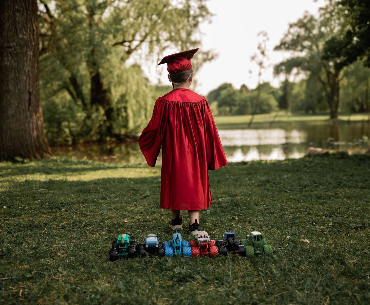 A graduate in red cap and gown stands by a pond with a collection of toy cars arranged at their feet.