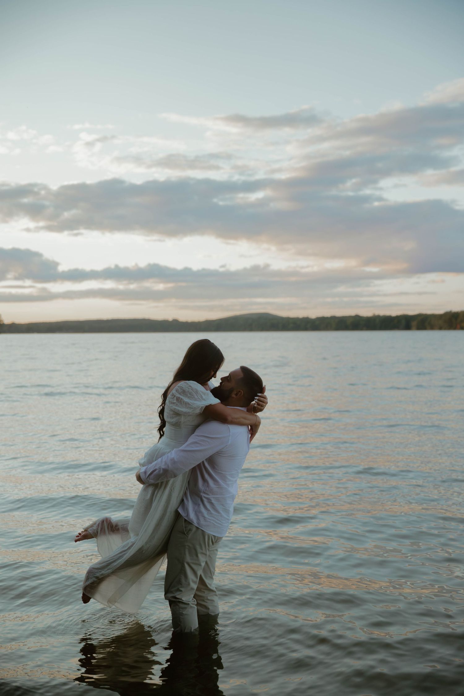 Couple shares romantic dance in ocean waters during sunset with coastal landscape behind them.
