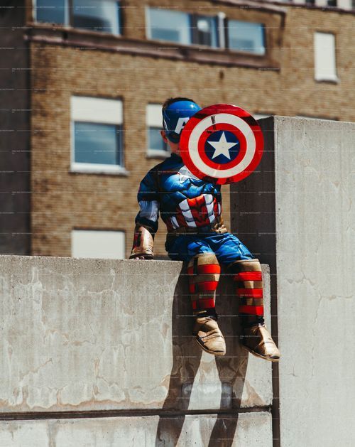 Captain America cosplayer sits on concrete ledge holding shield against brick building.