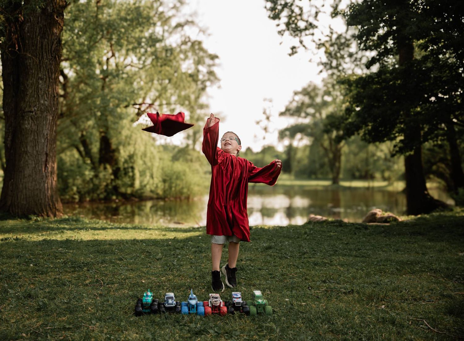 Red-robed graduate tossing mortarboard in celebration with toys arranged on grass by pond.