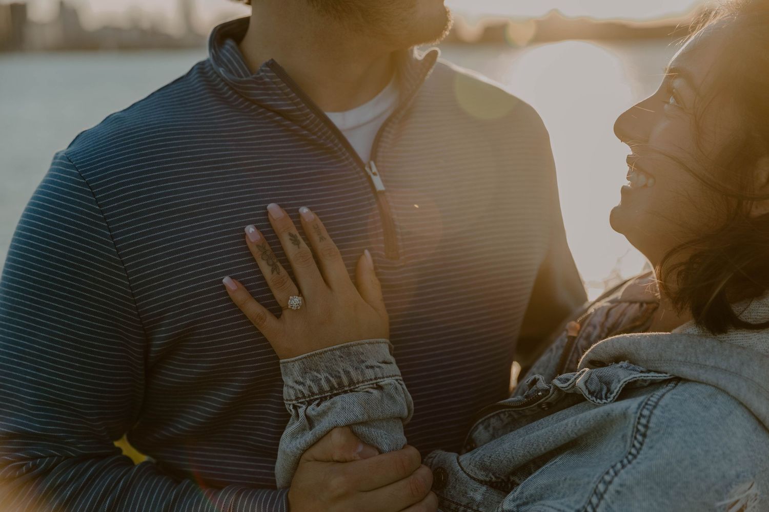 Couple embracing at sunset by the water, woman showing engagement ring, golden backlight glowing warmly.