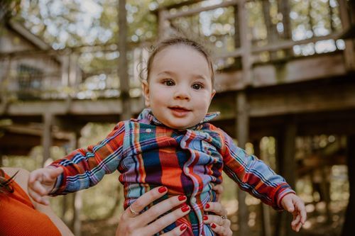 Happy toddler in plaid shirt reaching up while being held outdoors.