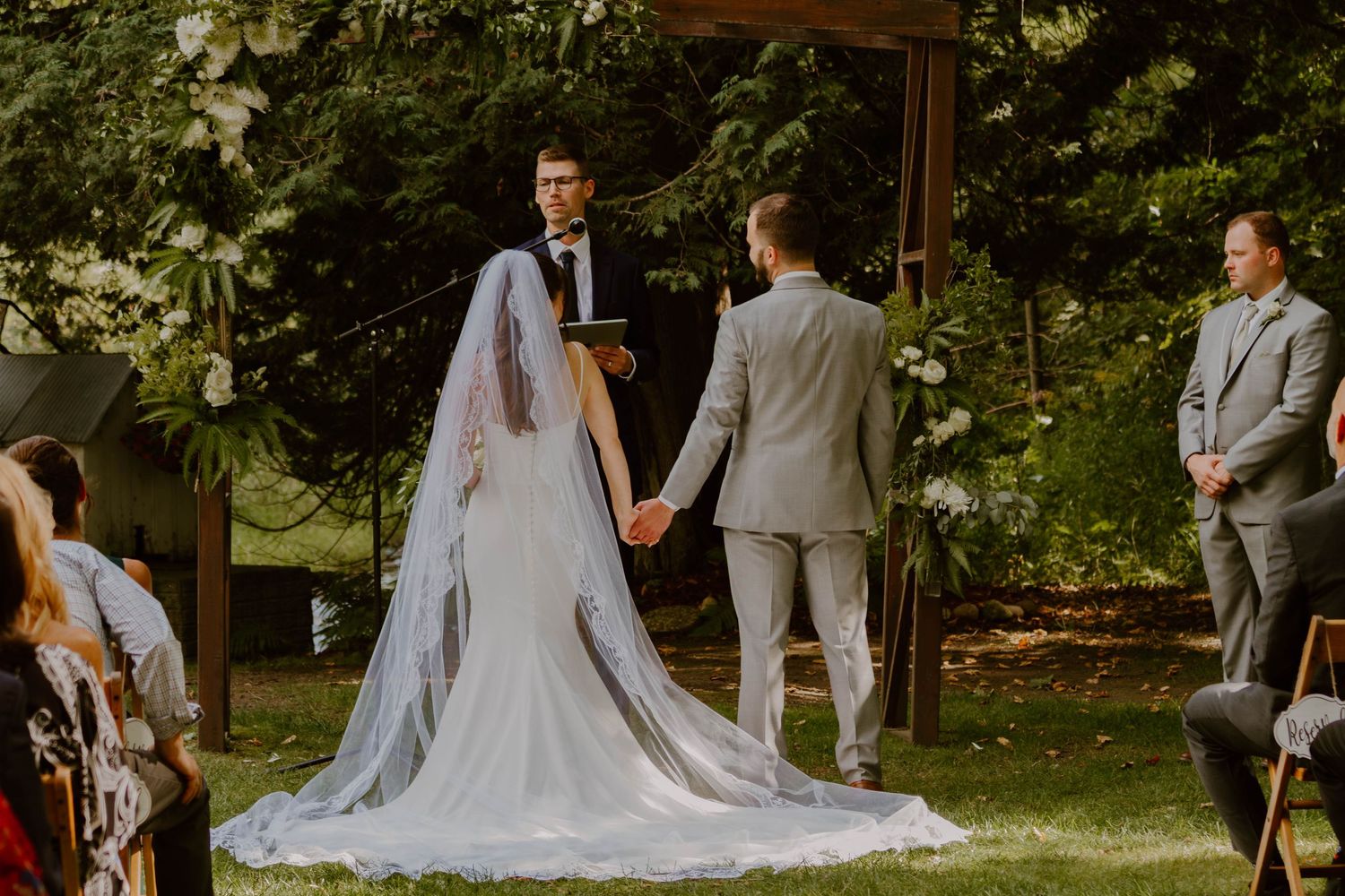 Outdoor wedding ceremony under trees with bride in flowing white gown and veil.