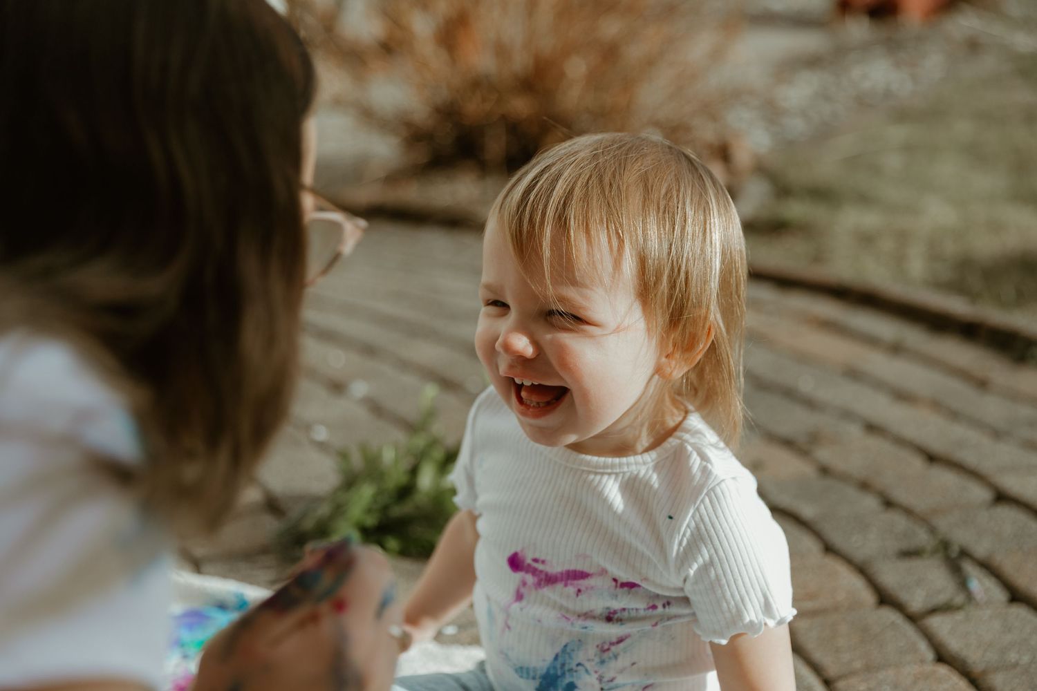 A joyful toddler laughs while sitting on a brick patio during a sunny day, wearing a white t-shirt.