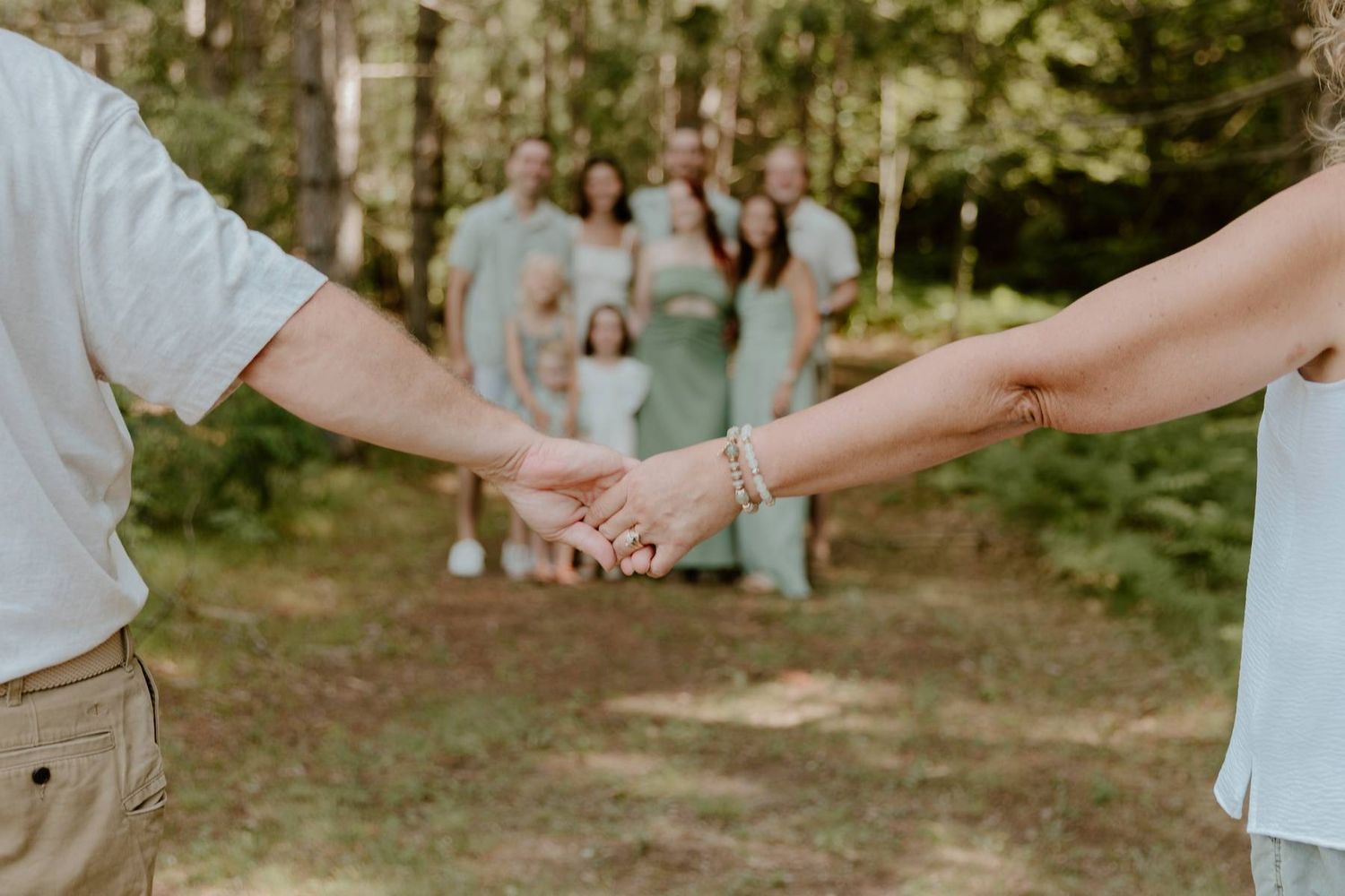 Couple holding hands in foreground with family group posed among trees in soft green tones behind them.