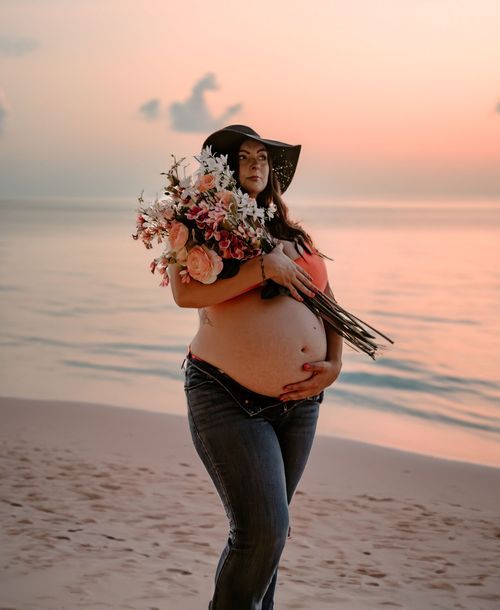 Beautiful maternity photoshoot at sunset on the beach with a large bouquet of flowers and wide-brimmed hat.