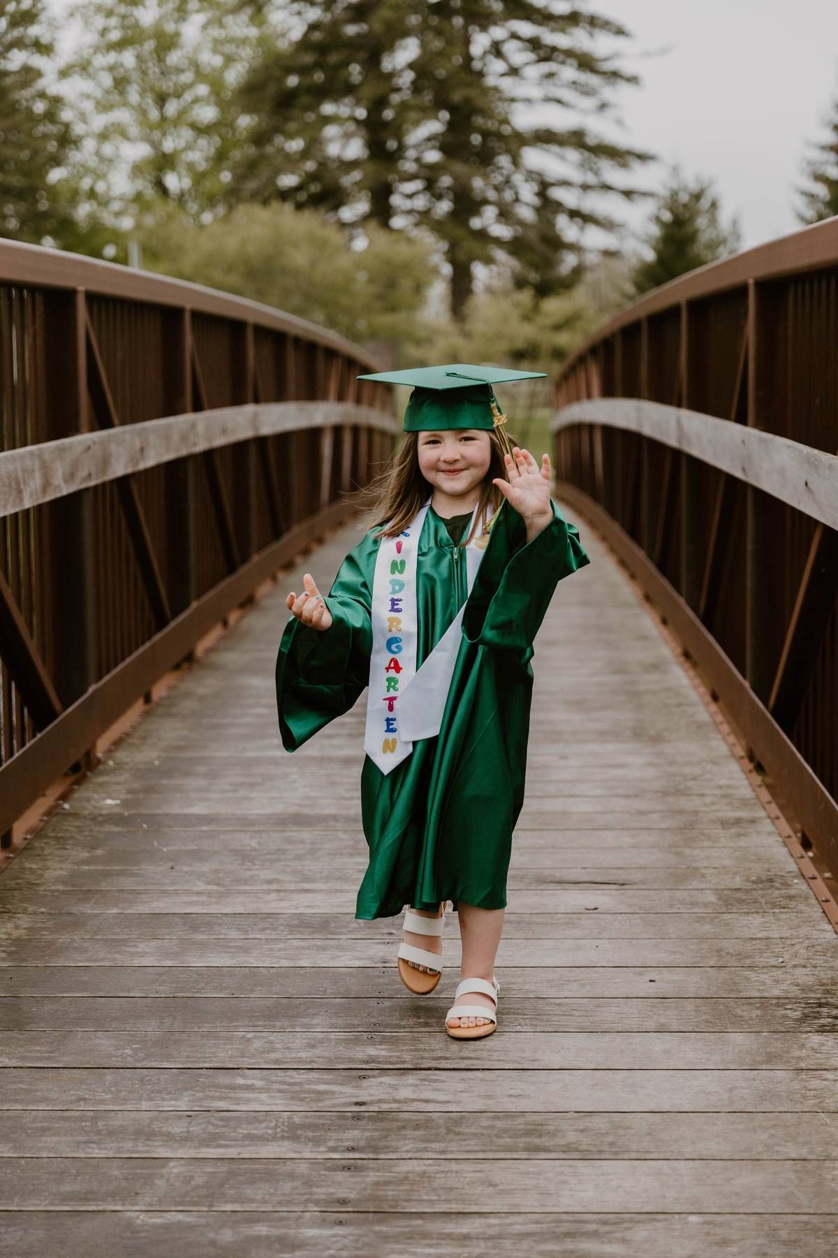Graduate in emerald green cap and gown walking on wooden bridge in natural outdoor setting.