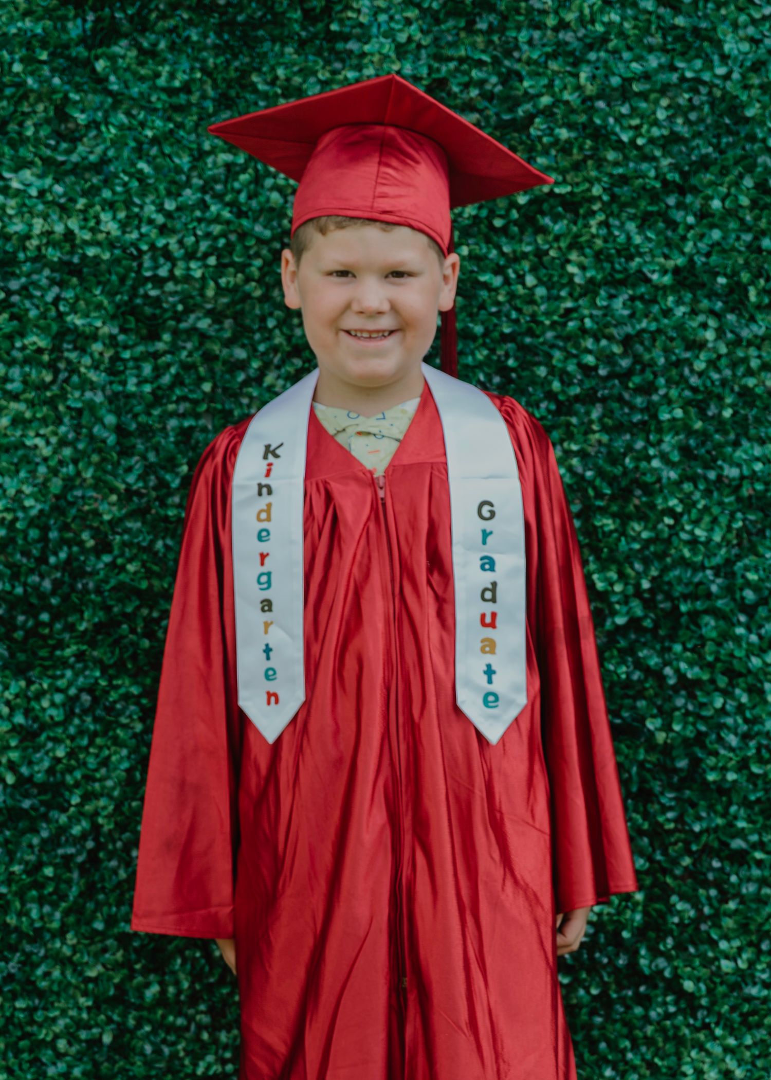 Young boy in red kindergarten graduation cap and gown with white sash, smiling in front of green ivy wall.