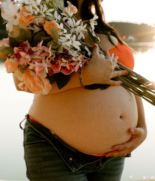 Close-up of pregnant belly with vibrant flower bouquet against beach backdrop at sunset.