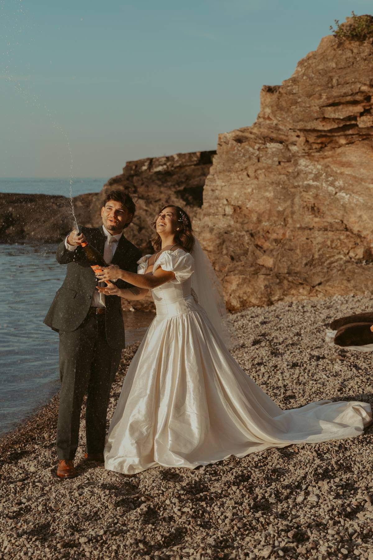 A bride and groom celebrate on a rocky beach, popping champagne beside a cliff at sunset in wedding attire.