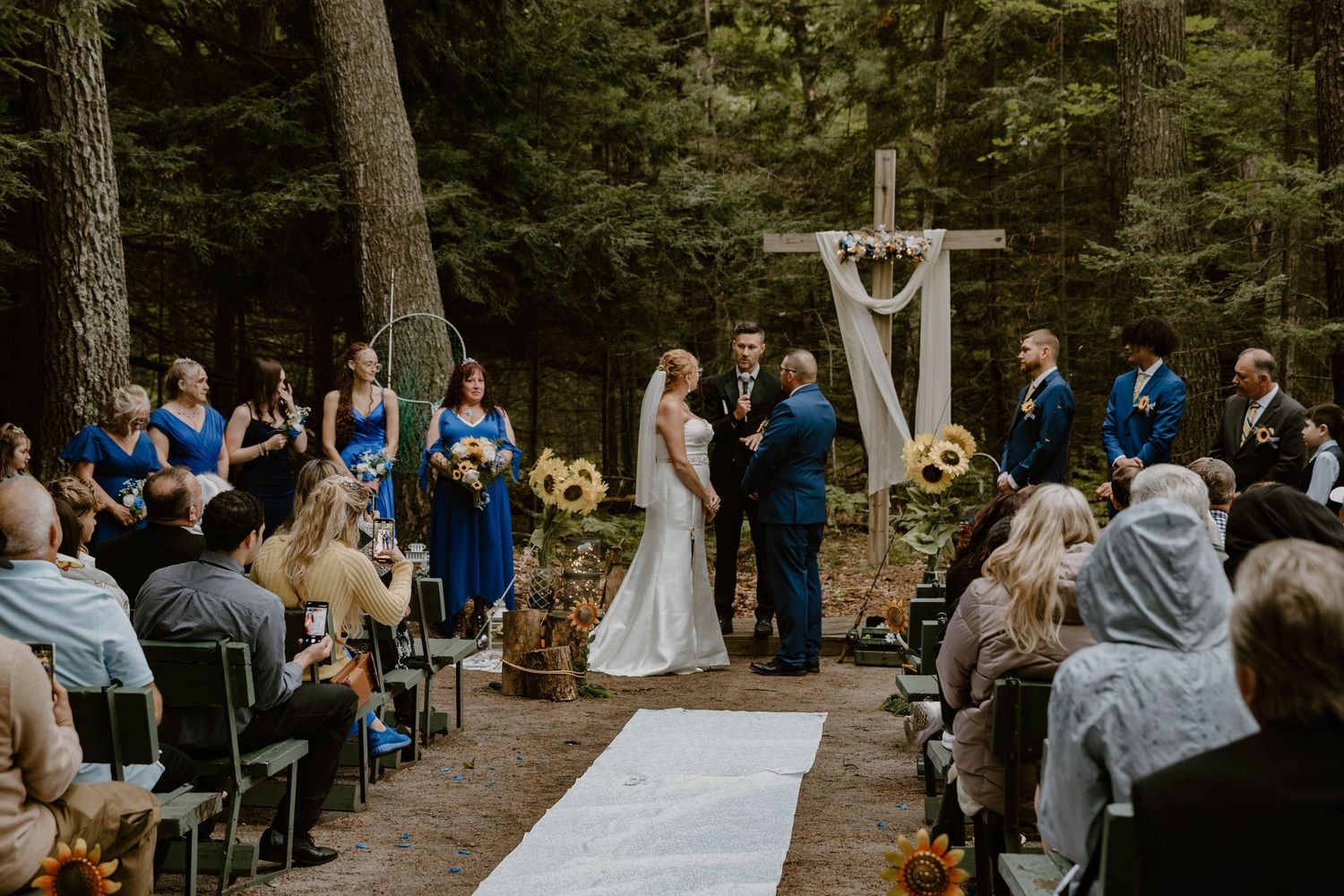 Outdoor forest wedding ceremony with white arch and aisle runner surrounded by guests in blue attire.