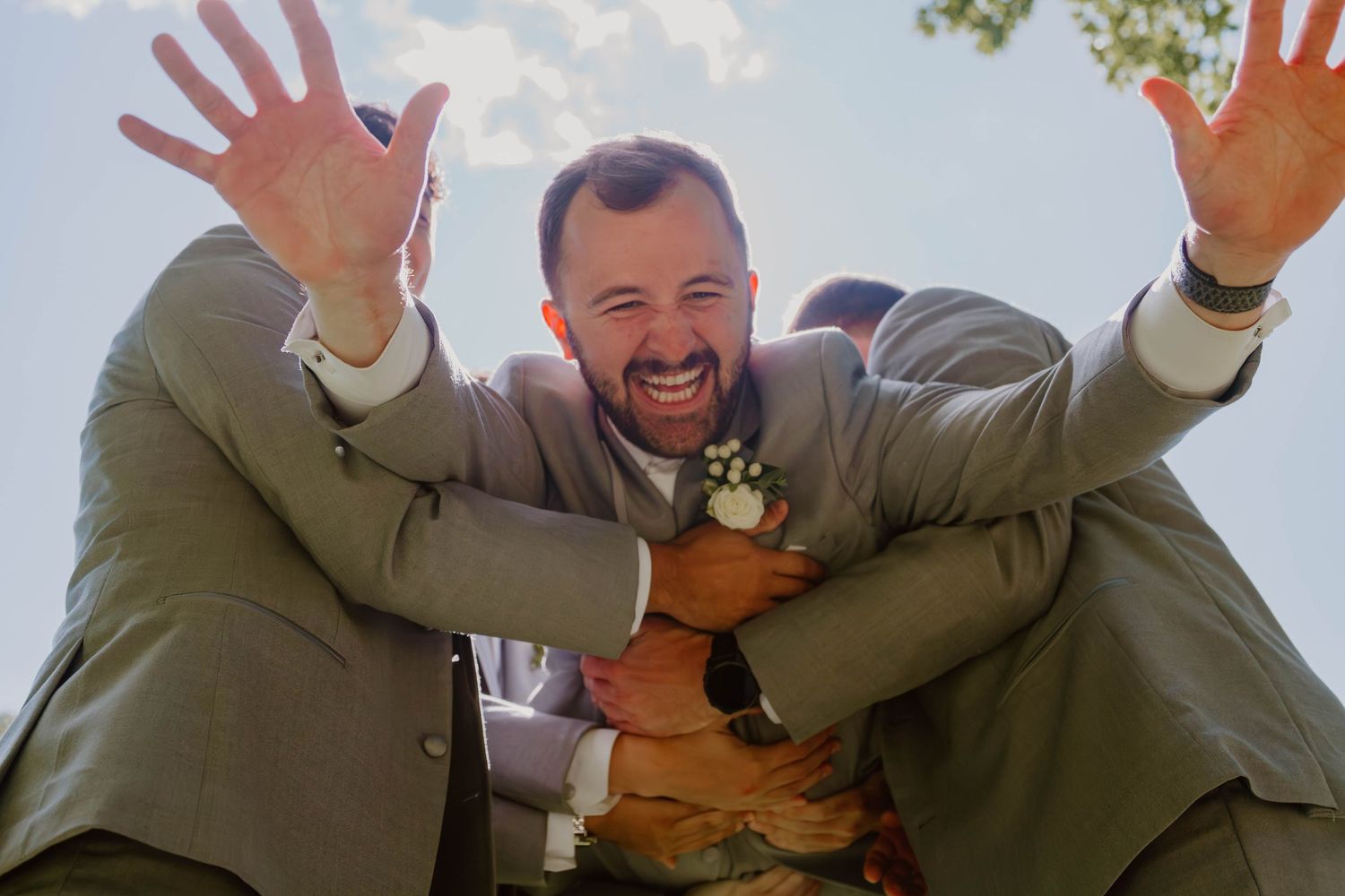 A group of people in olive colored jackets wave and smile enthusiastically against a bright sky background.
