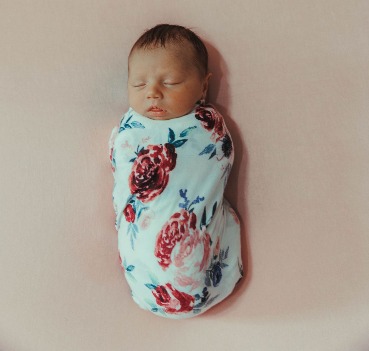 Sleeping newborn swaddled in mint green floral print blanket lying against neutral beige background.