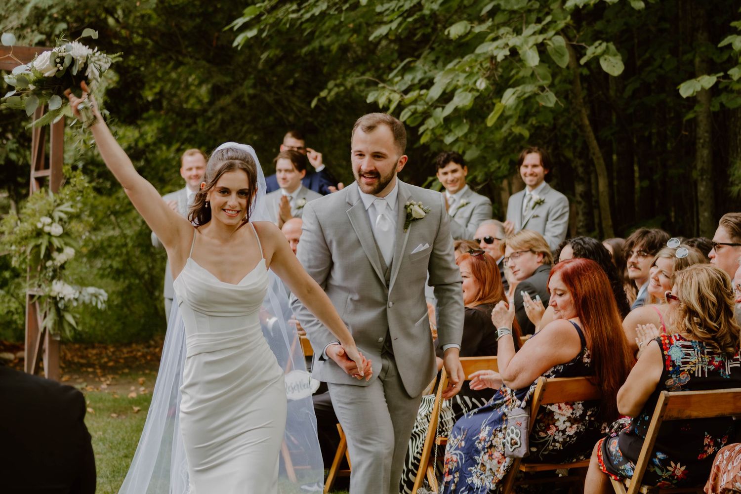 Newlyweds celebrate with raised arms while walking down the aisle at an outdoor forest wedding ceremony.