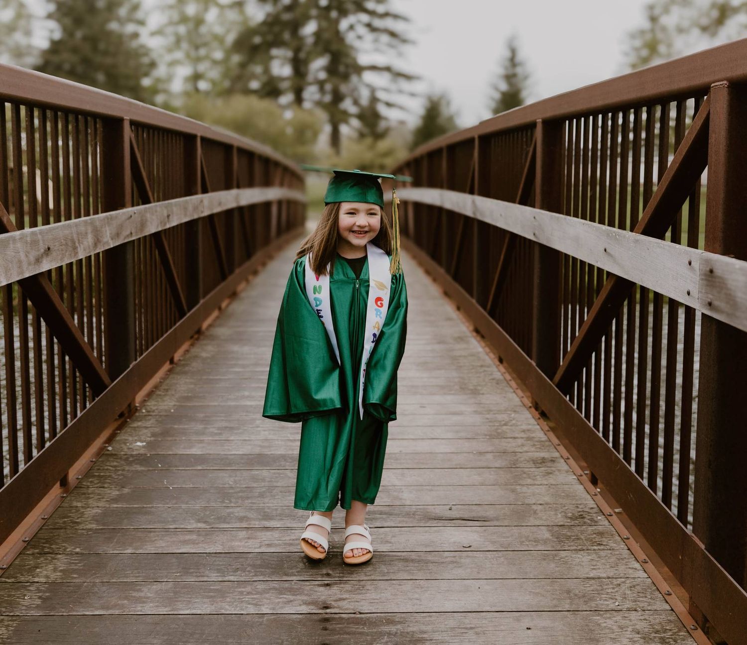 A person in a green graduation gown walks across a wooden bridge surrounded by trees on a sunny day.
