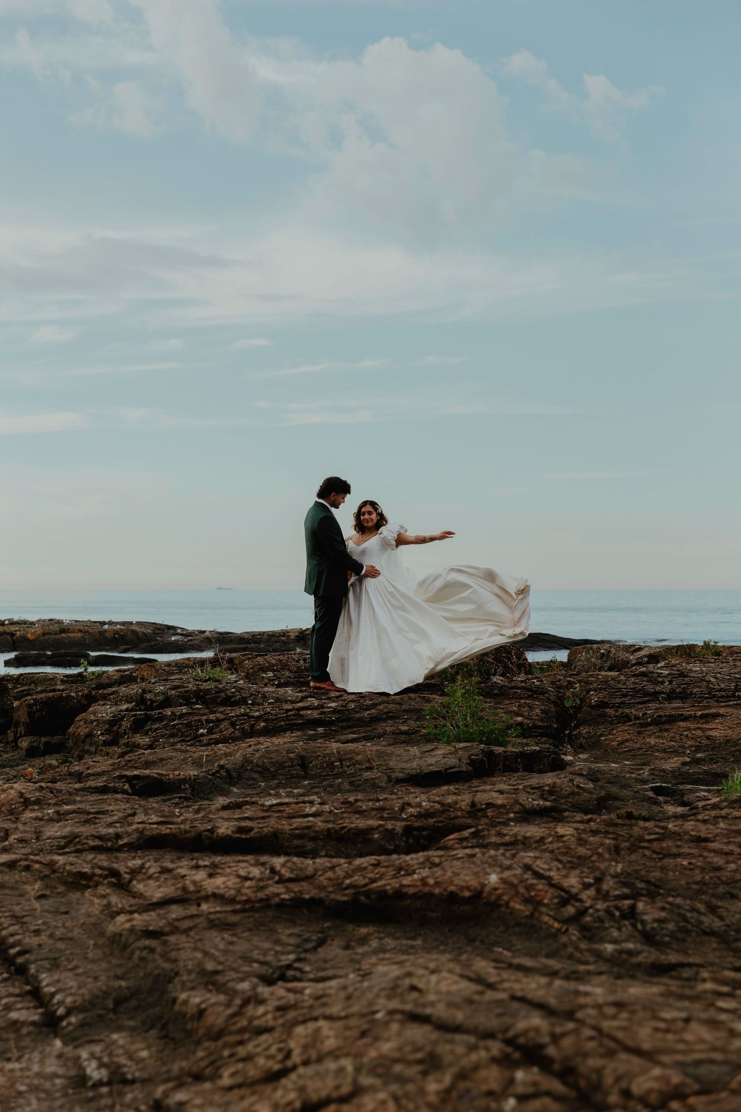 Bride and groom stand on rocky coastal cliffs, her white gown flowing in the wind, with ocean and blue sky behind them.