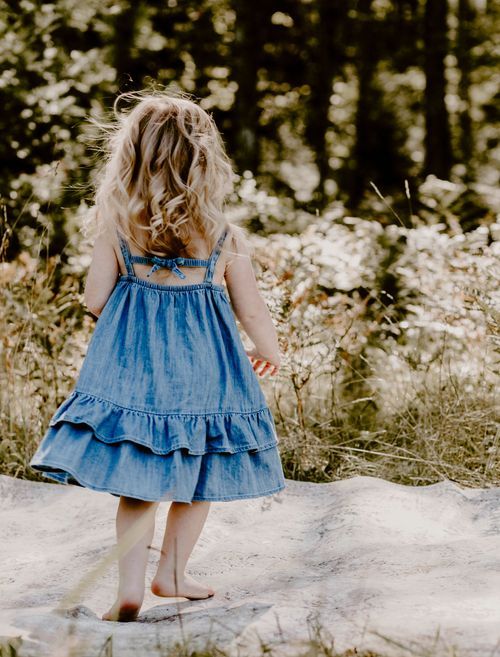 View from behind of a child in a blue ruffled dress standing on light colored ground.