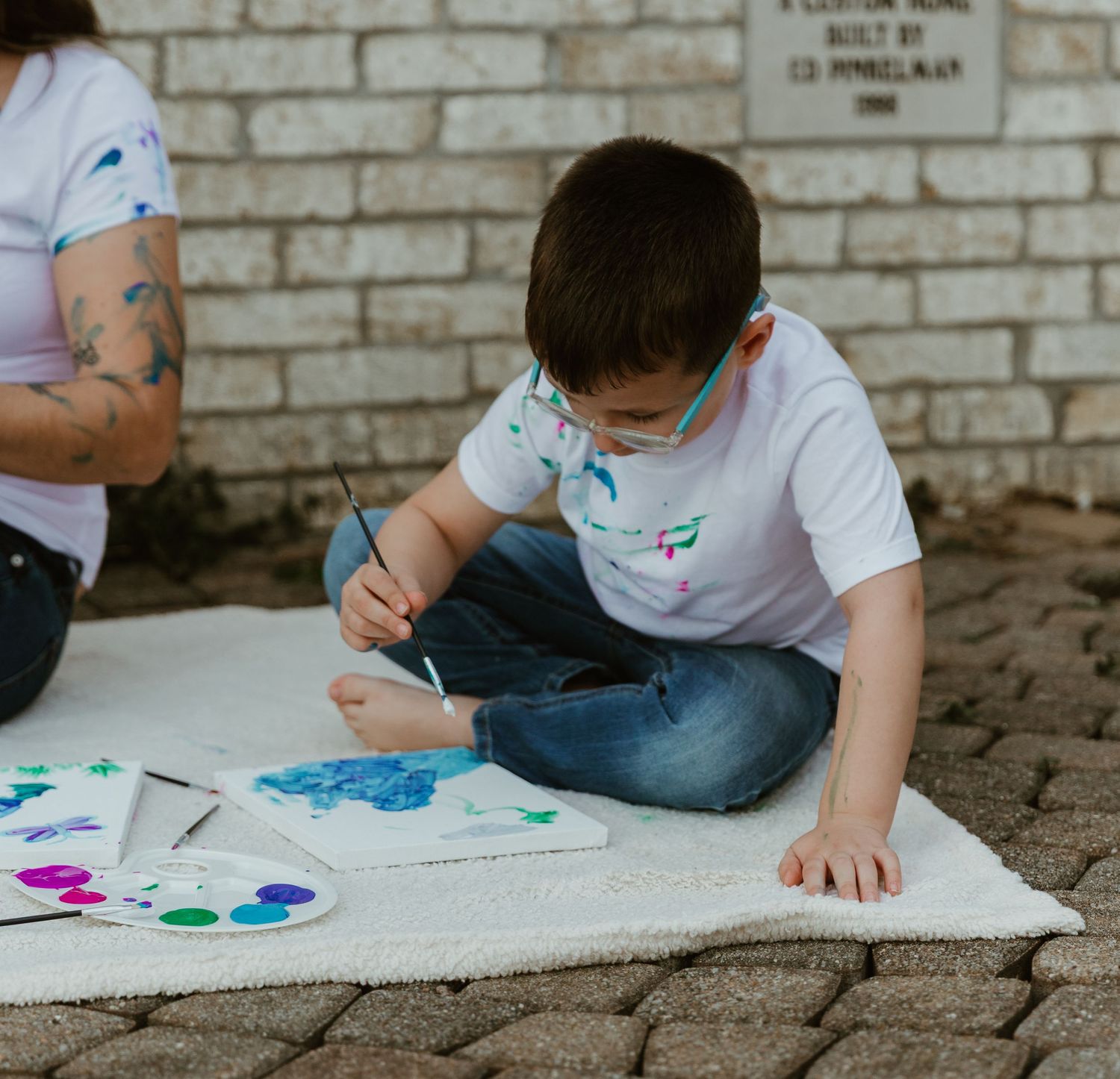 Child in white polo shirt sitting on concrete and painting on a white canvas with colorful paints.