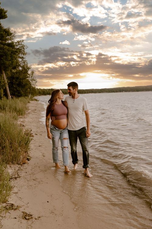A couple strolls barefoot along a sandy beach at sunset with dramatic clouds over a lake.