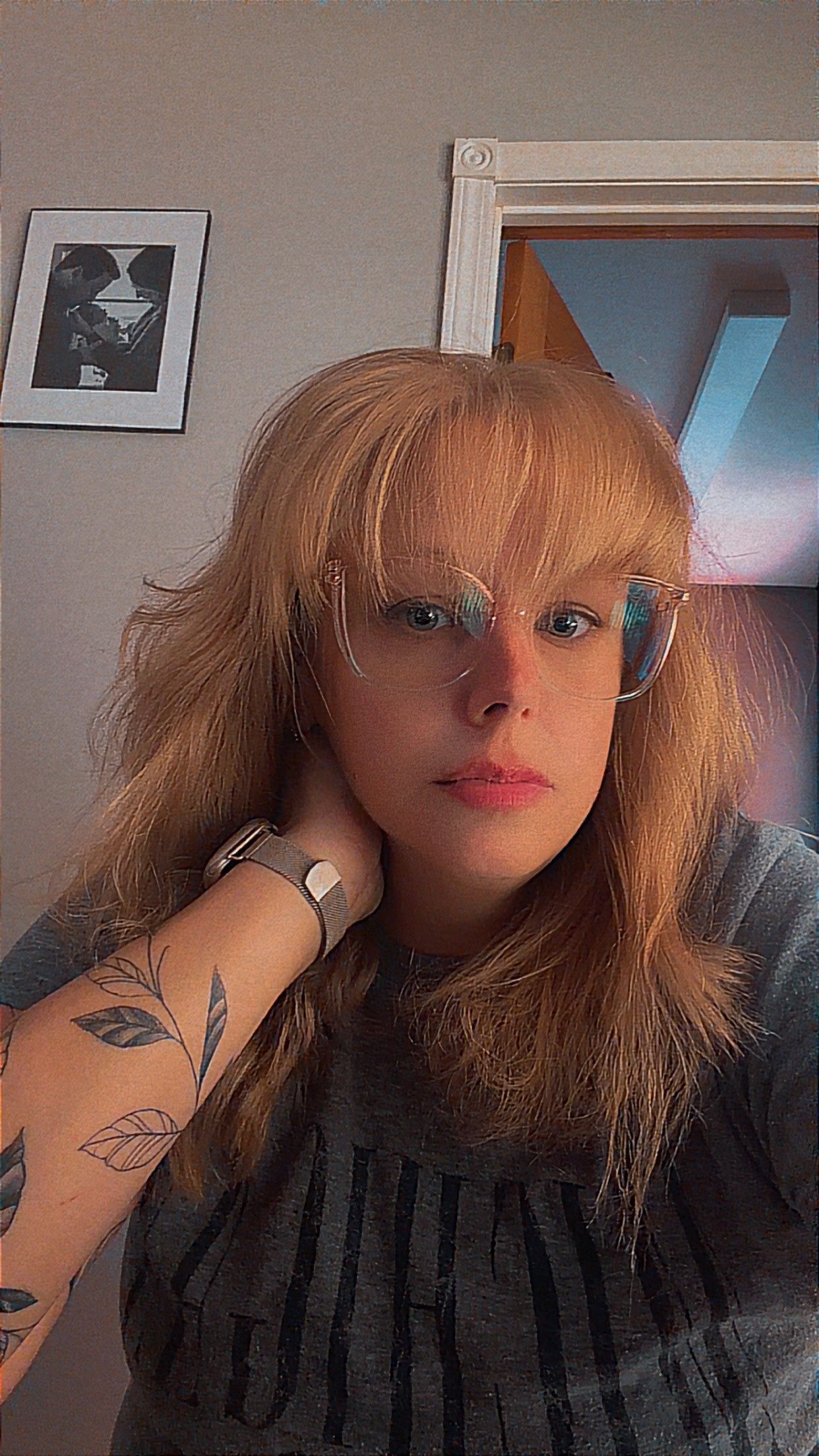 Young woman with blonde wavy hair and clear glasses, wearing a gray tee, showing leaf tattoos on her arm.