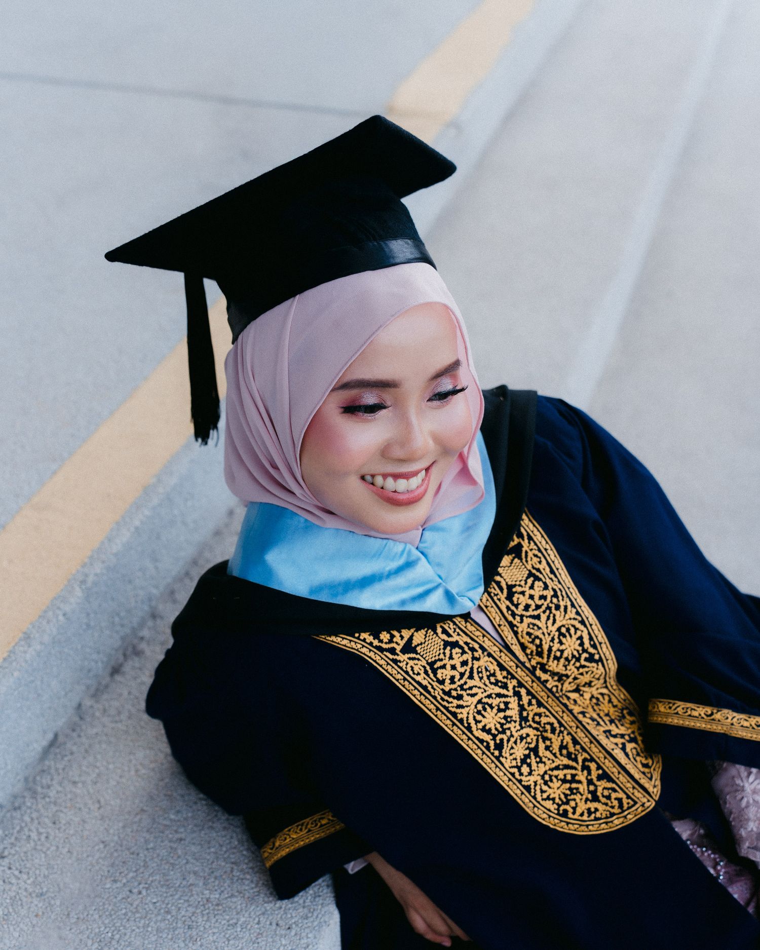 Graduate in black academic regalia with gold embroidery and light blue scarf smiles while sitting on stairs.