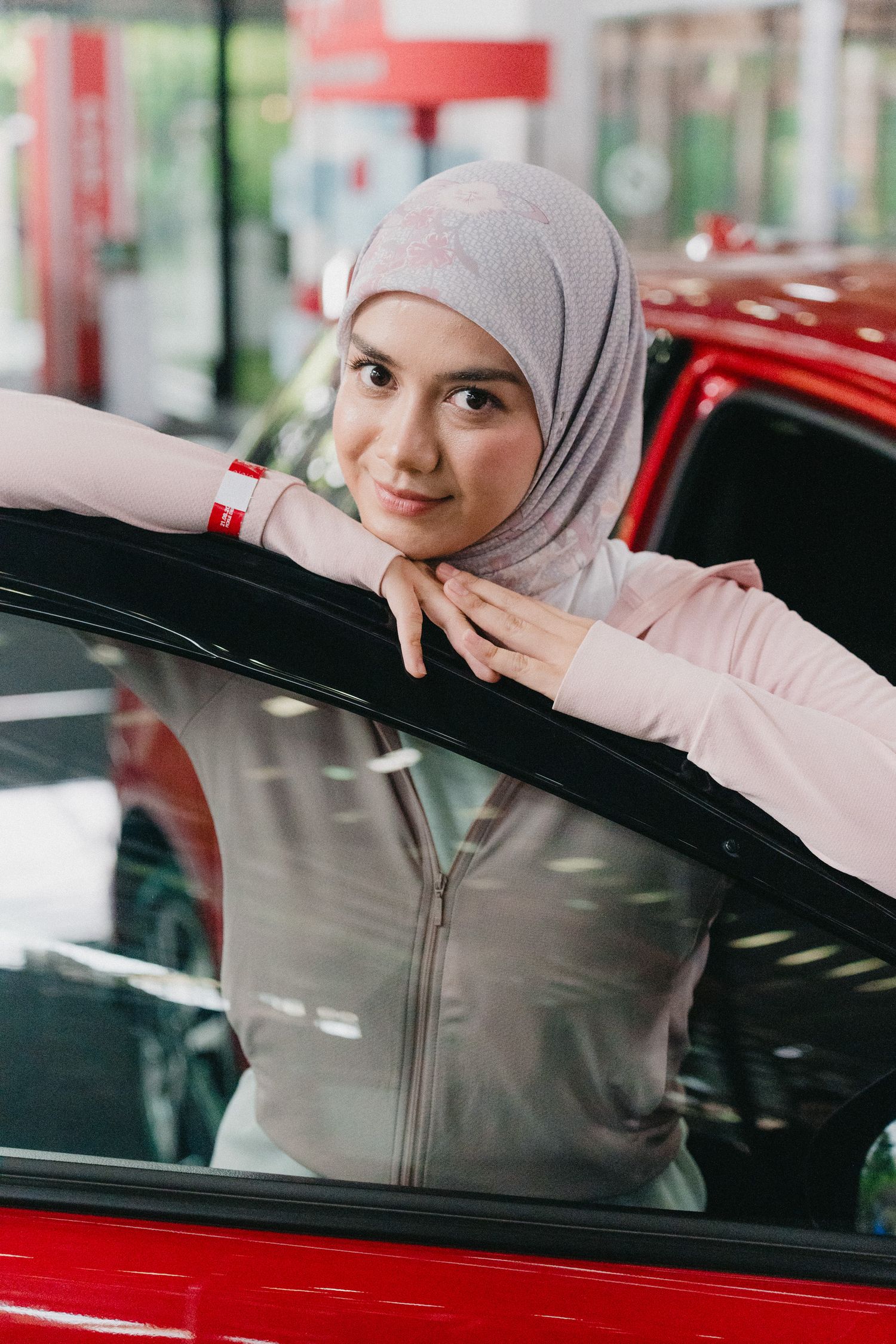 A woman in a light gray hijab and athletic wear leans on a red car door with a gentle smile.