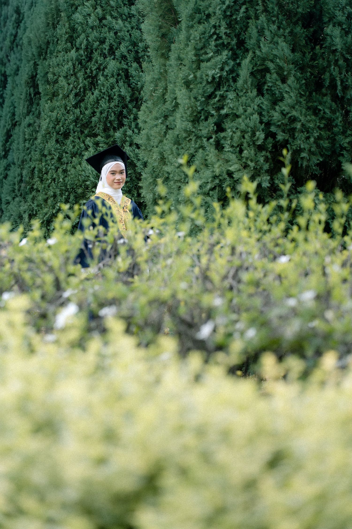 A person in white hijab stands in a field of wildflowers with evergreen trees in the background.