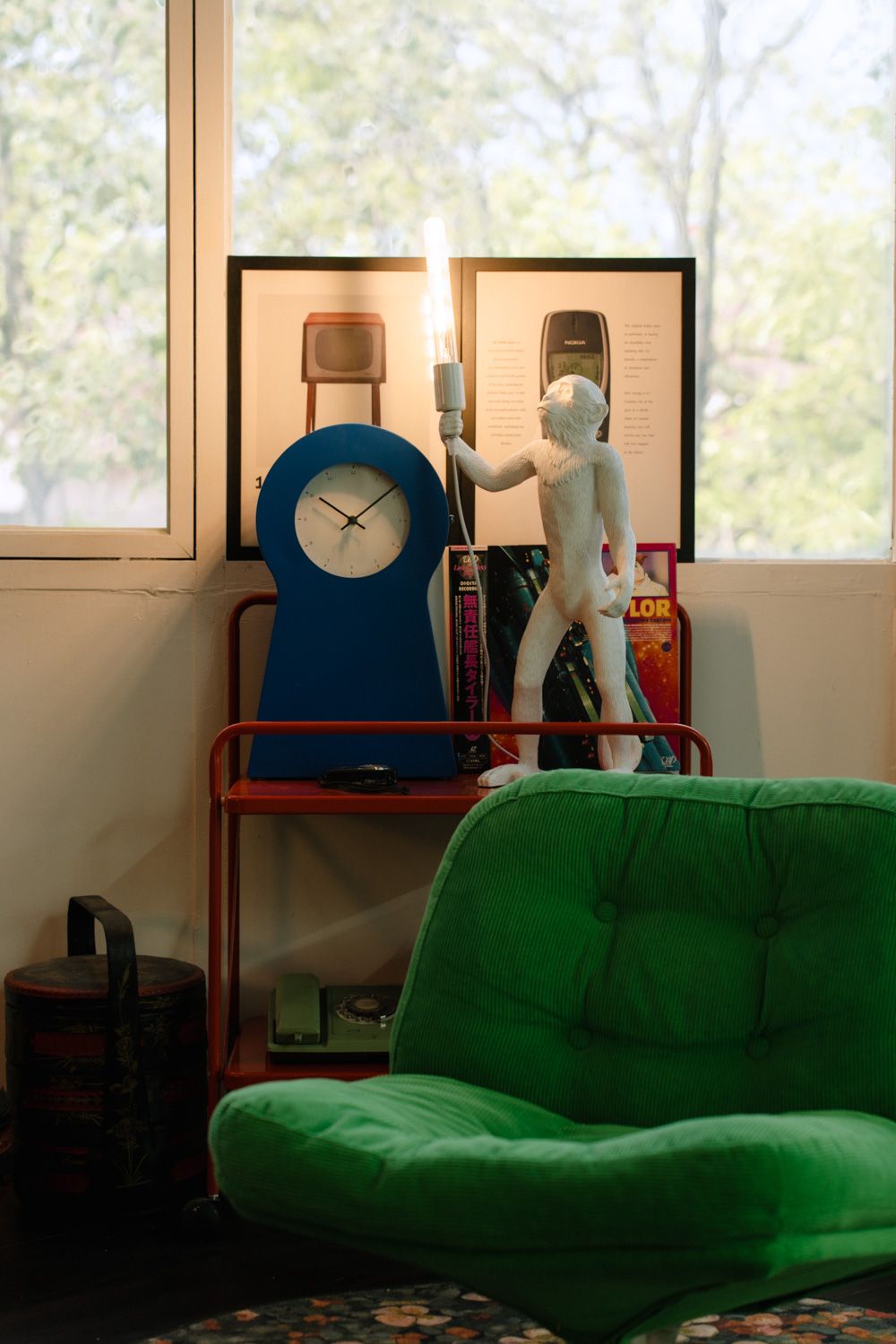 Retro room with green velvet chair, blue clock, white figurine, red shelf, and vintage phone poster by sunlit window.