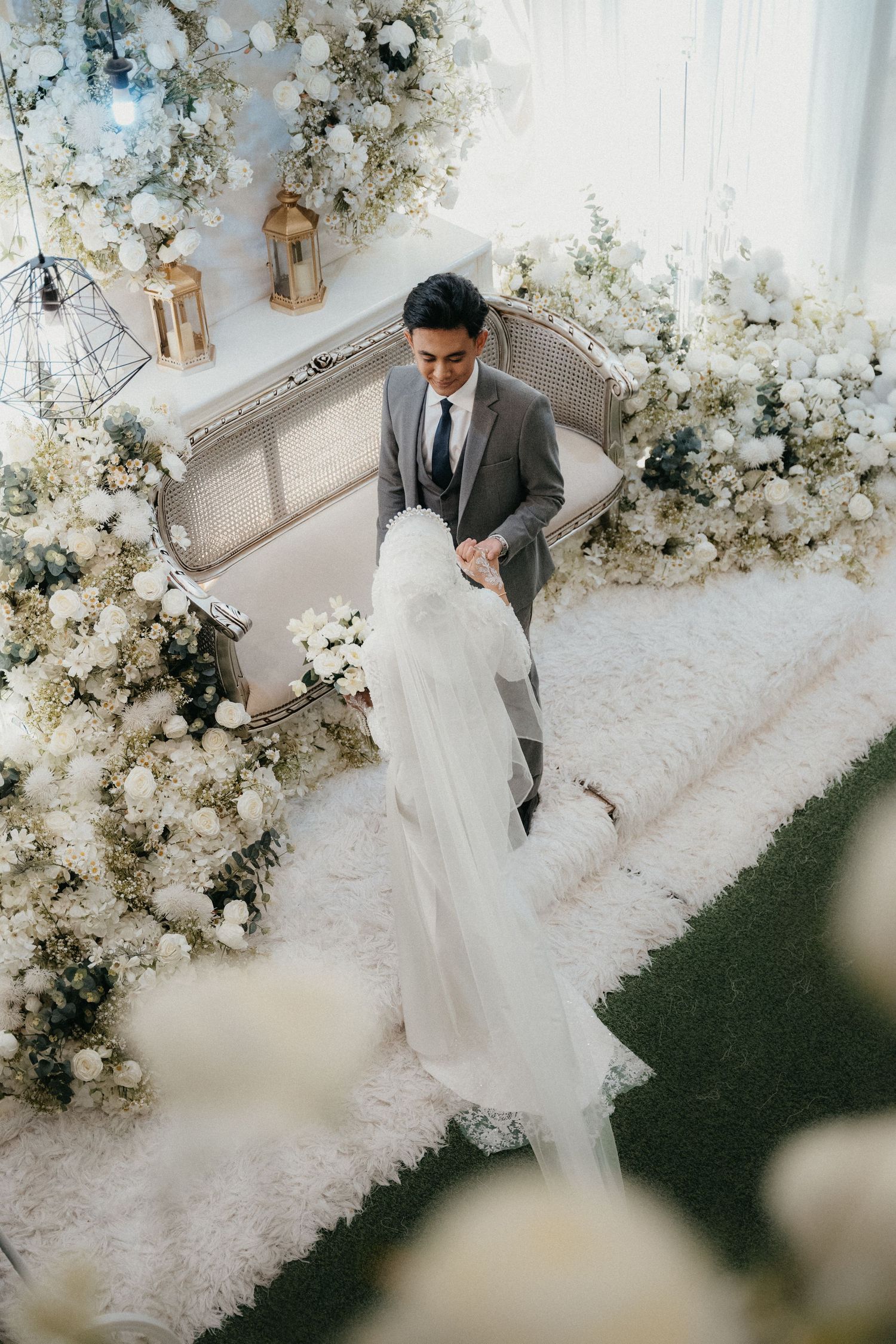 A wedding scene decorated with luxurious white floral arrangements and lanterns as the couple stands on a light carpet.