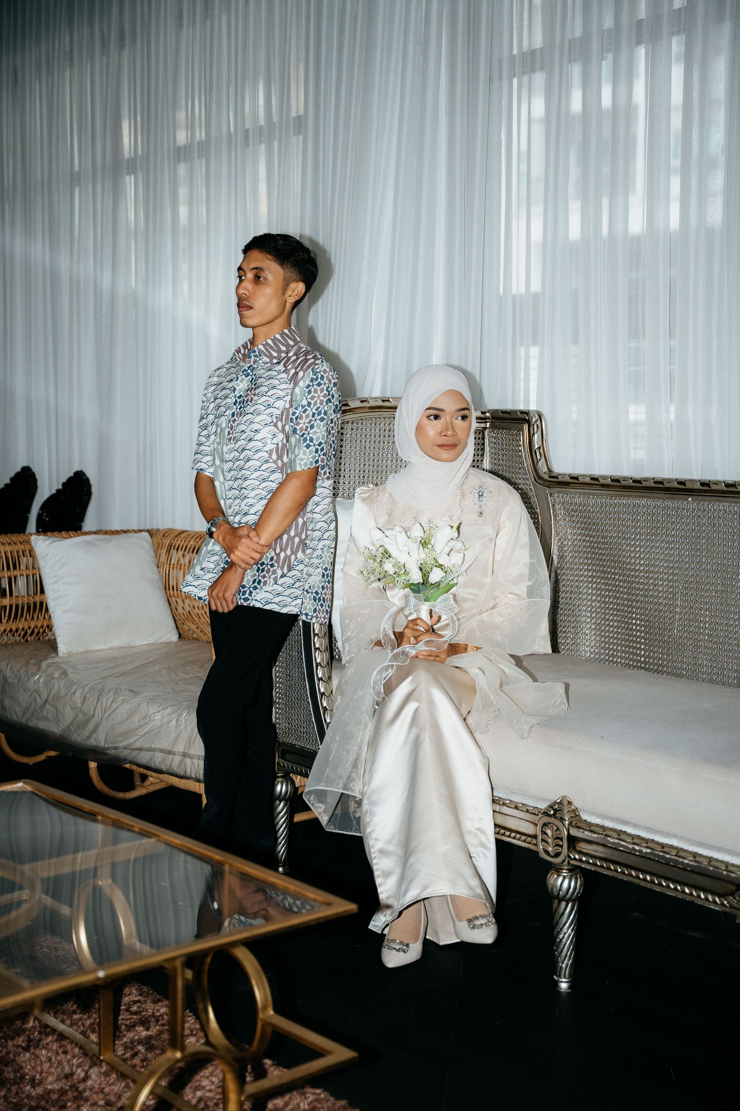 Two elegantly dressed people in a modern living room with sheer white curtains and vintage furniture.