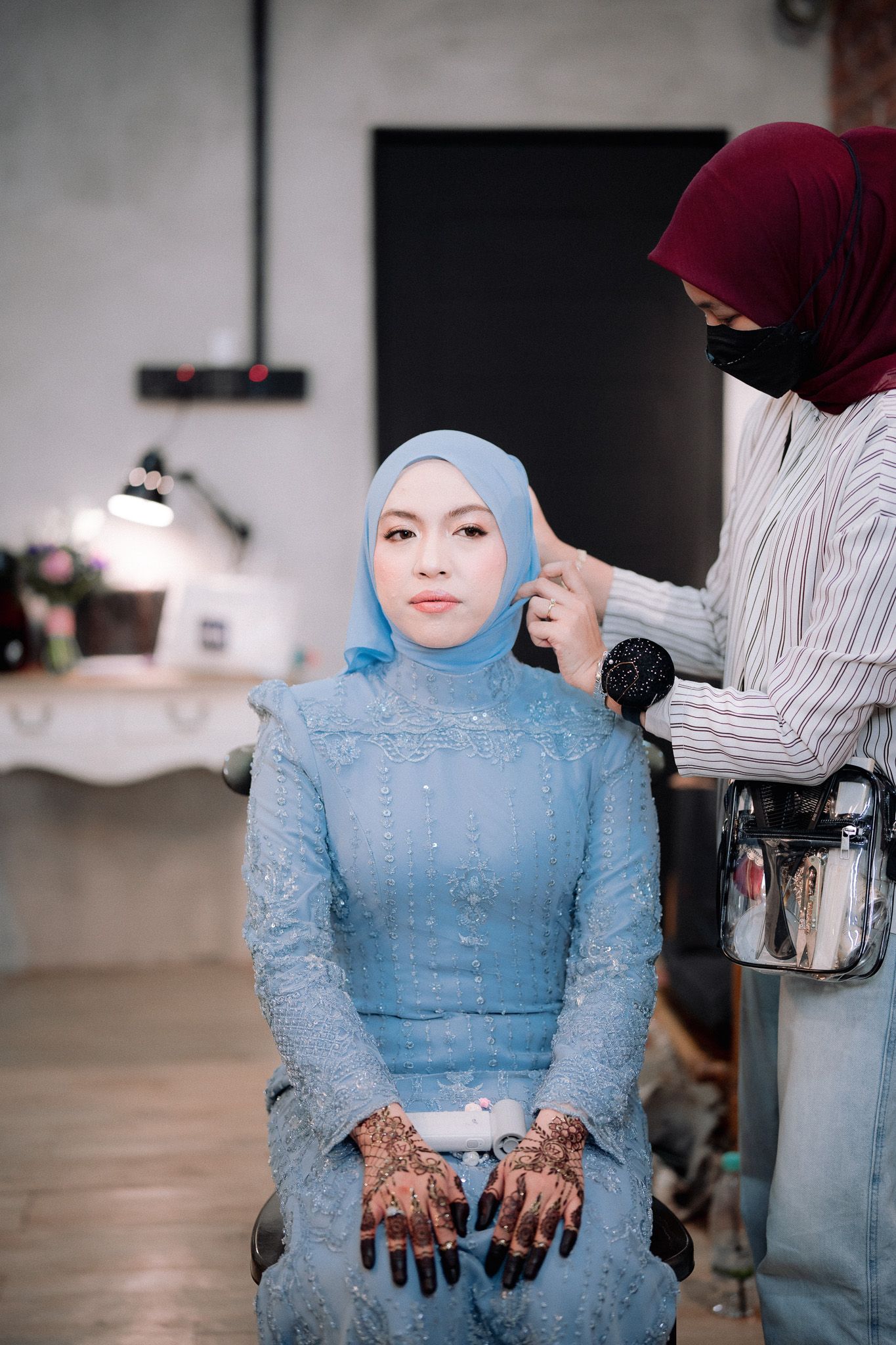 A bride in blue hijab and kebaya gets makeup done by artist in maroon hijab, with henna on her hands.