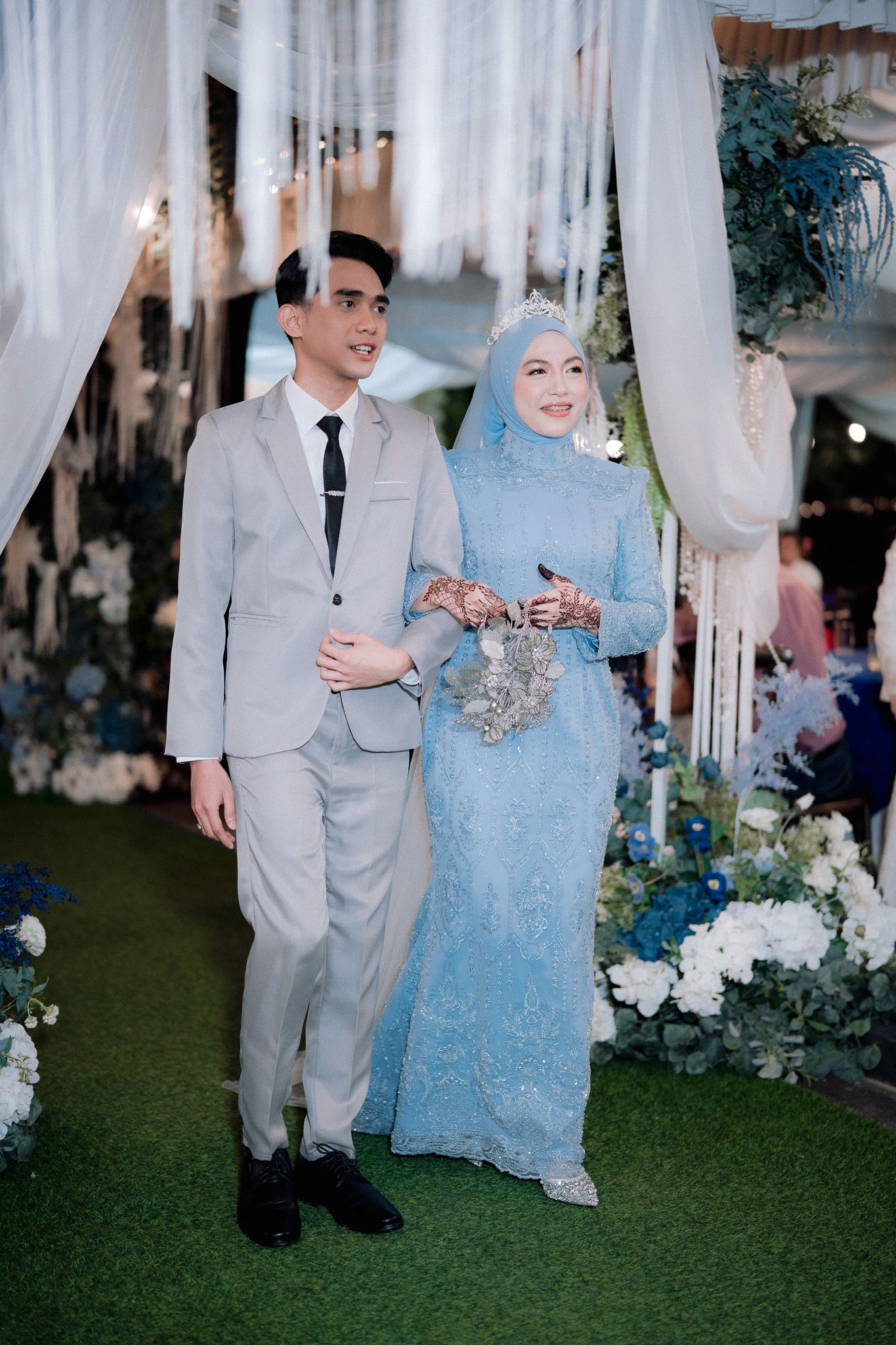 Muslim bride in light blue hijab gown and groom in gray suit walking together at elegant blue-themed wedding ceremony.