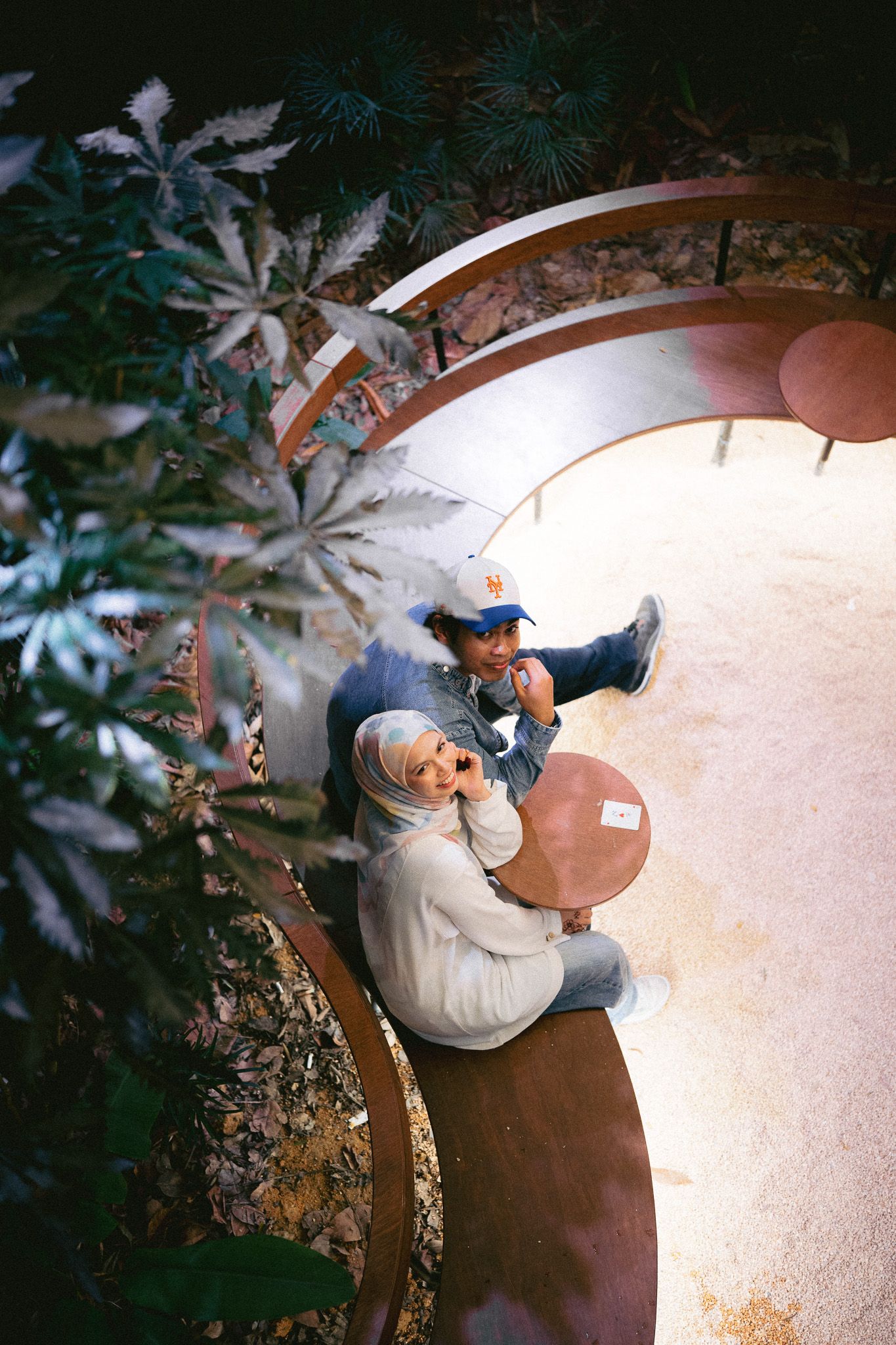 Aerial view of two people sitting on a curved bench surrounded by lush plants in an outdoor garden setting.