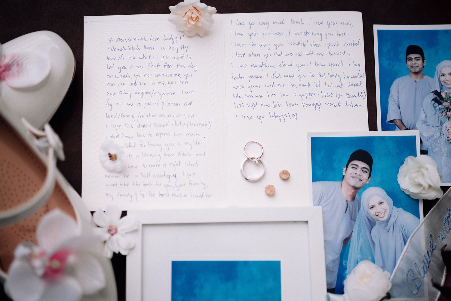 Wedding flat lay with handwritten vows, gold rings, earrings, photos of couple in blue outfits, and white flowers.