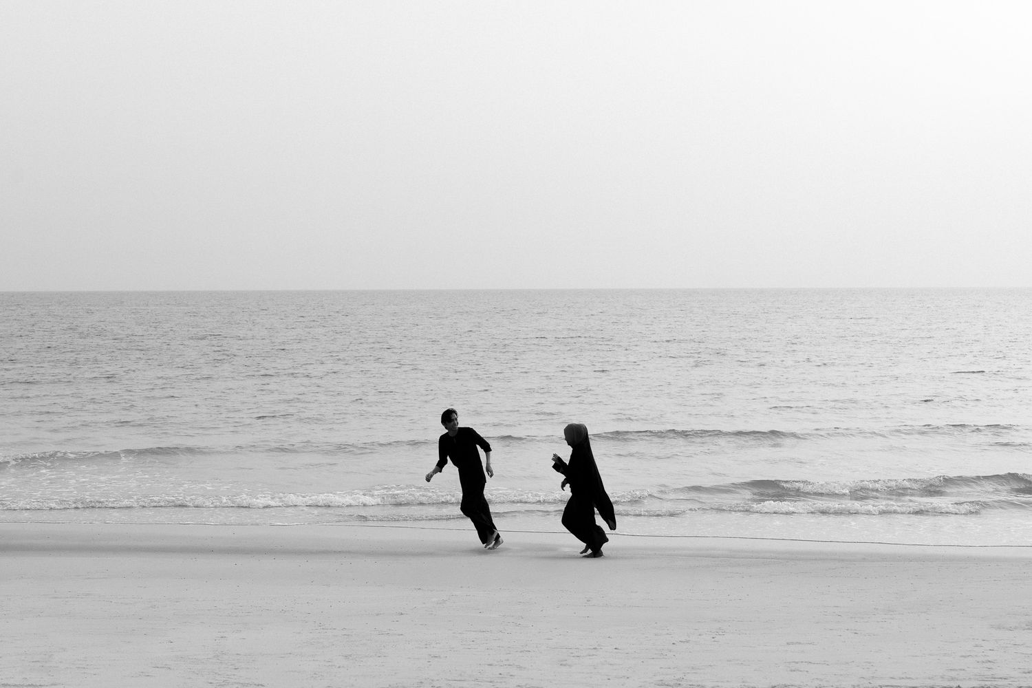 Two people in dark clothing walk along a serene beach shoreline in black and white photography.