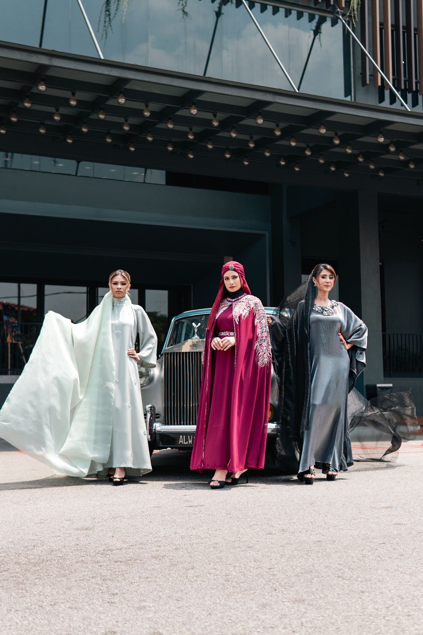 Three models in flowing modest dresses posing in front of classic Rolls Royce.