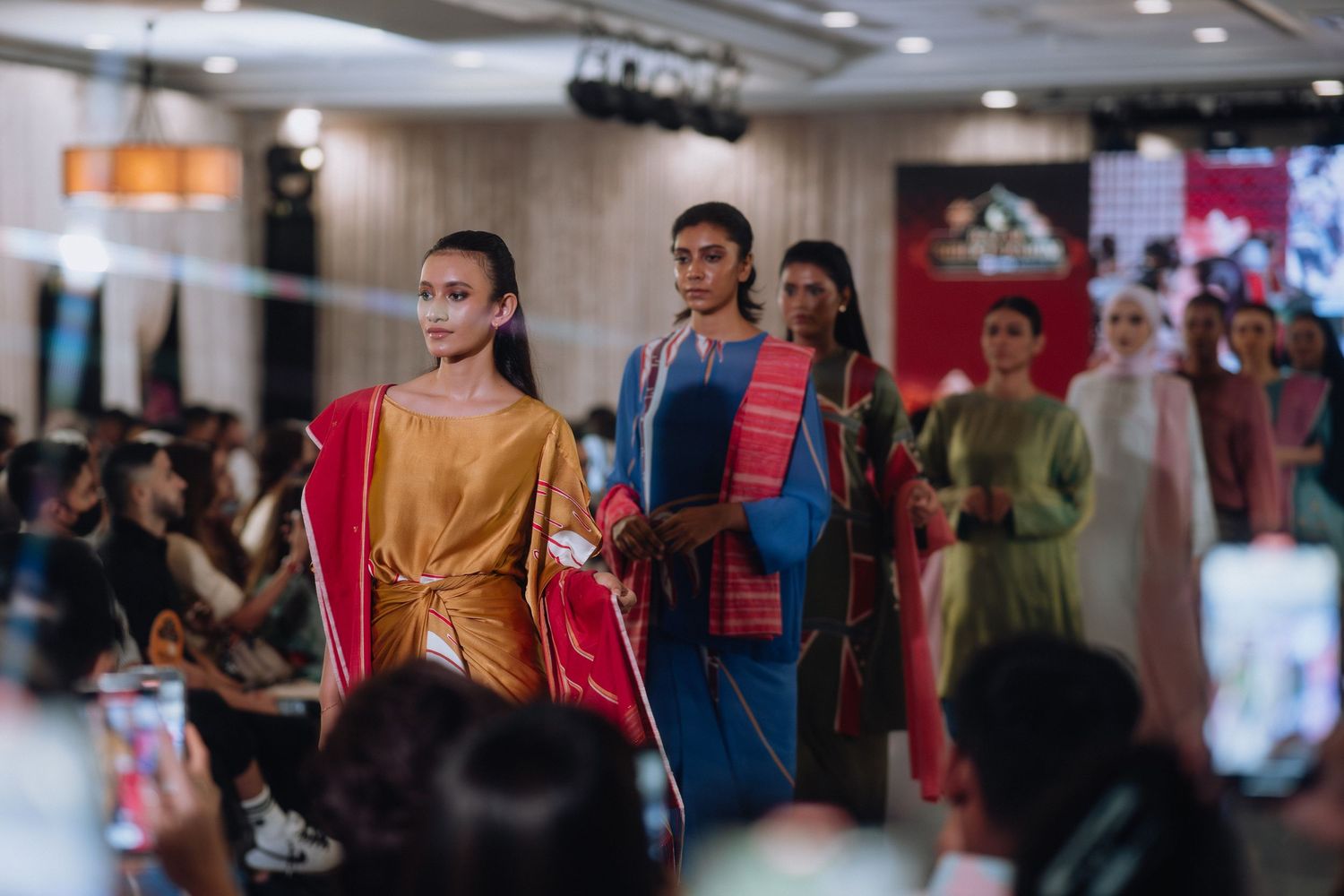 Models showcase colorful contemporary fashion designs on indoor catwalk with audience.