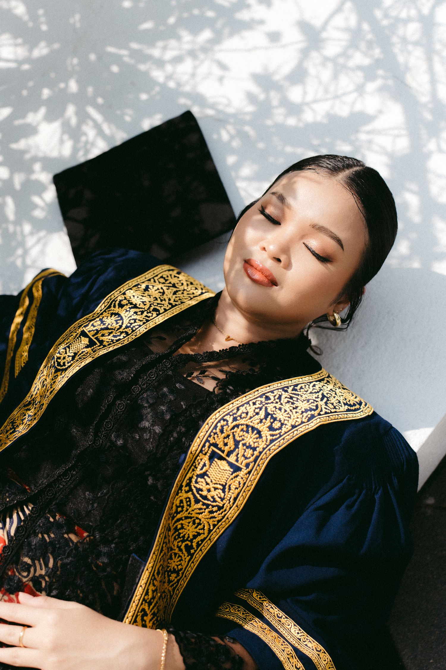A model poses in an elegant velvet graduation gown with ornate gold embroidered trim against a dappled light background.