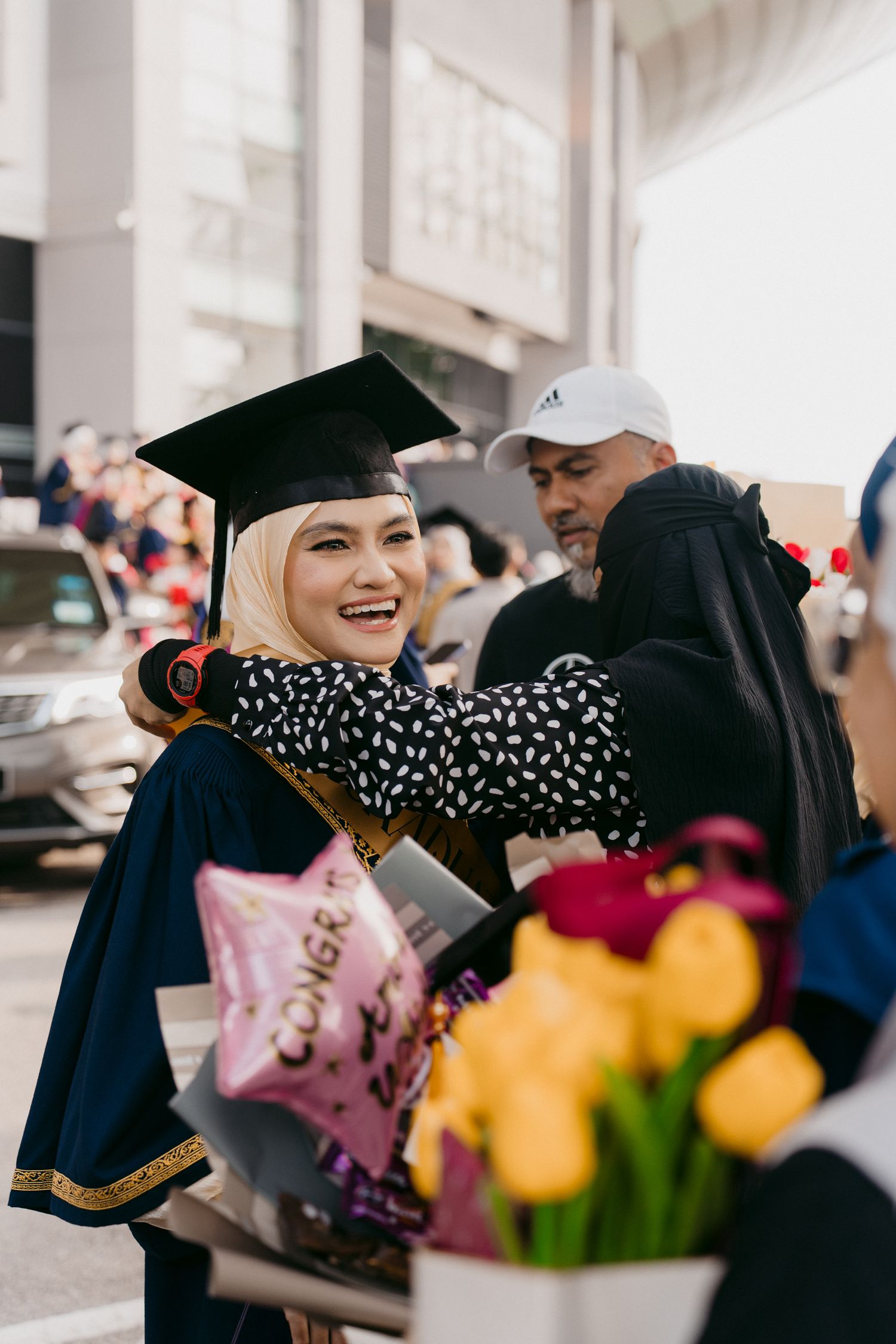 A graduate in cap and gown celebrates on graduation day with flowers and a congratulatory balloon nearby.