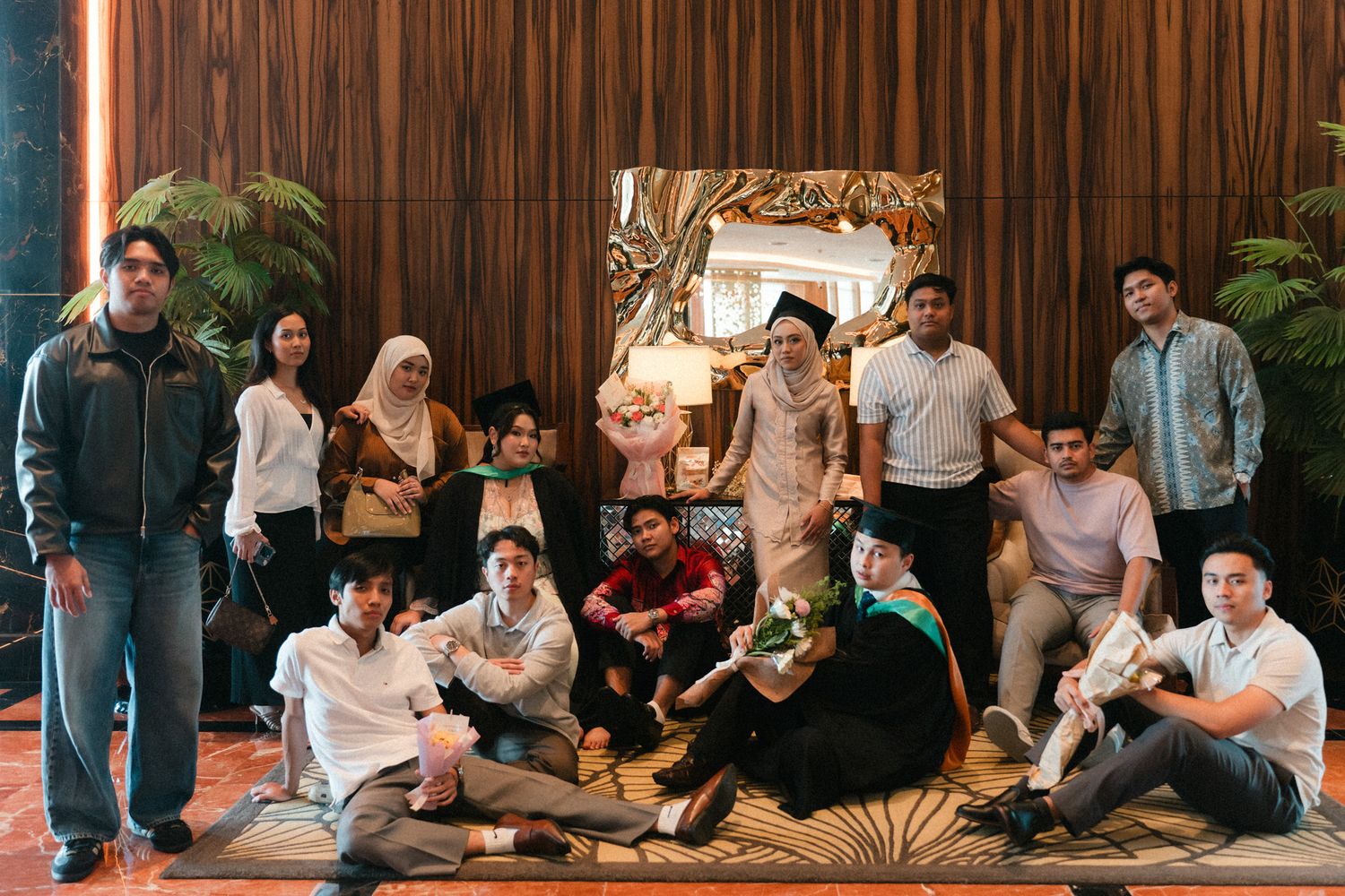Group of people posing casually together in front of a wooden wall with decorative frame and plants.