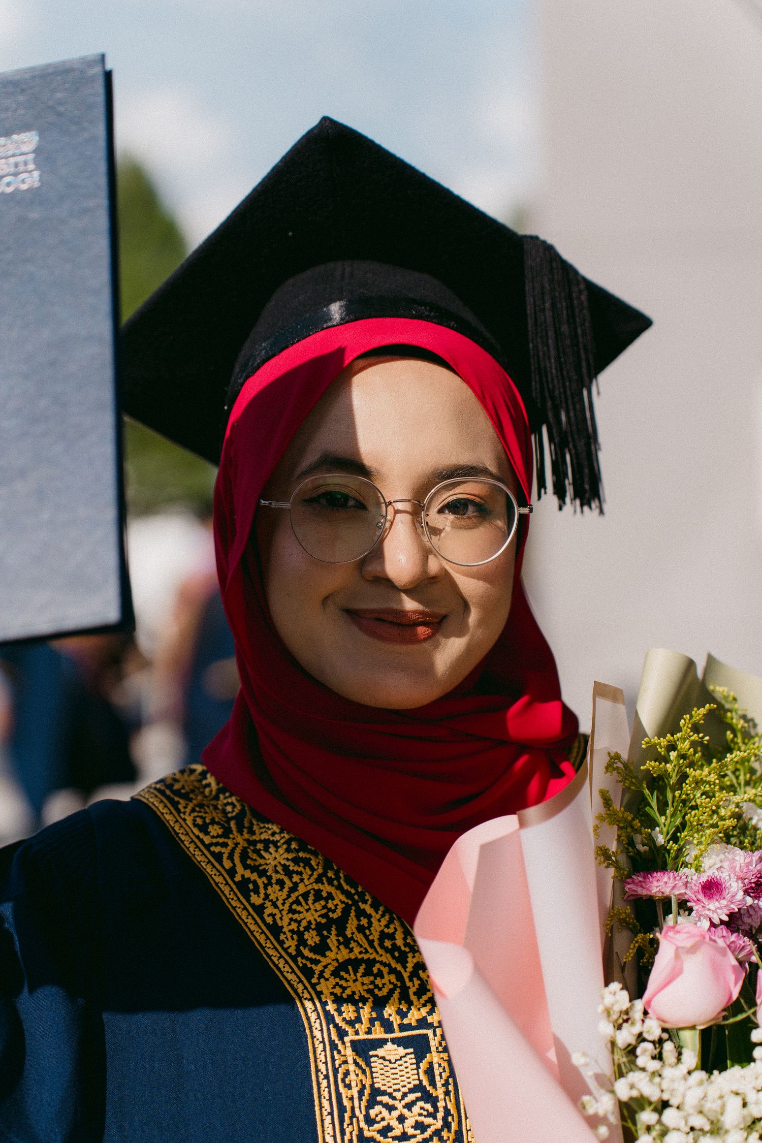 A graduate in glasses and red hijab poses with diploma and flowers while wearing traditional academic dress.