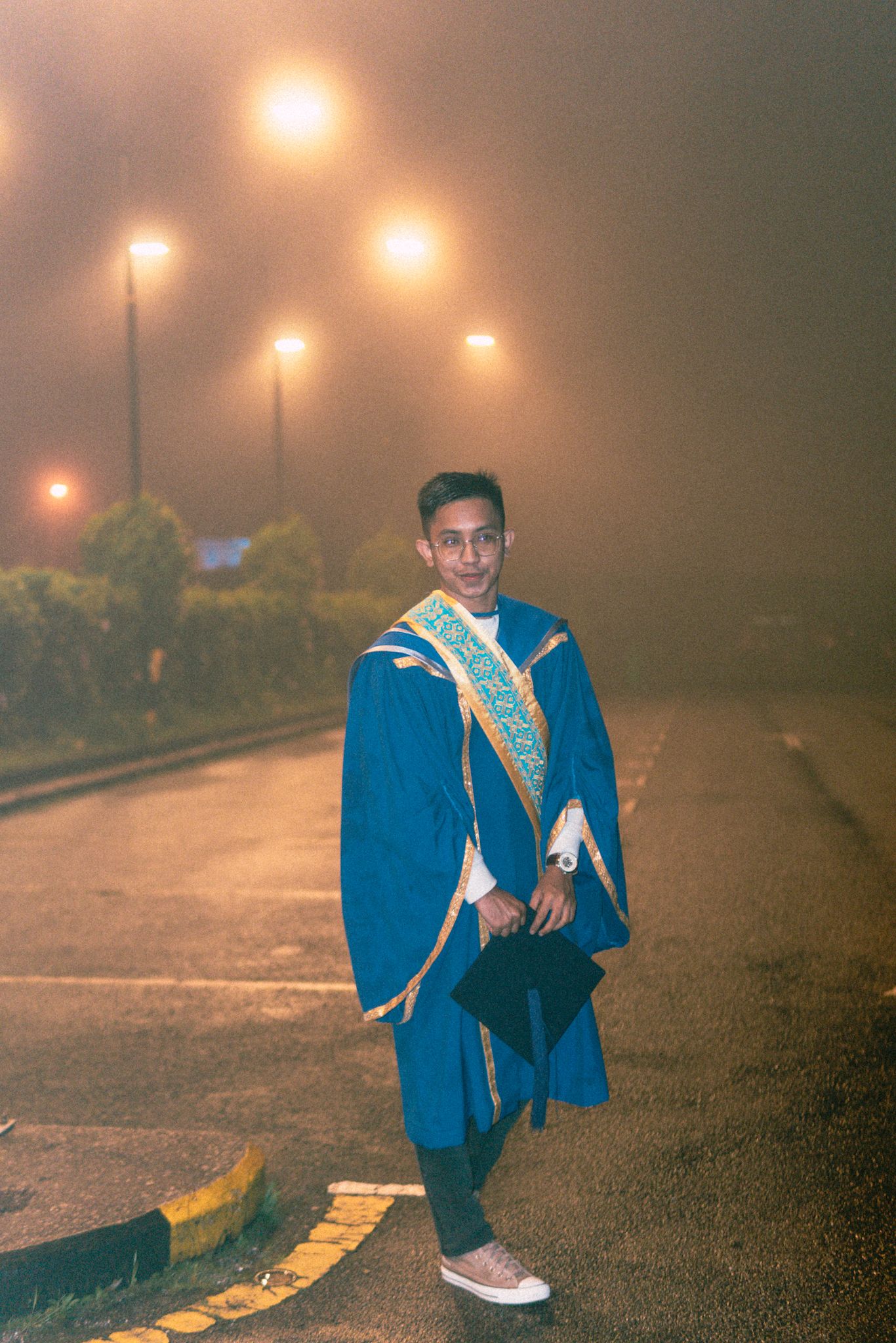 Graduate in blue academic regalia posing on foggy street with glowing streetlights.