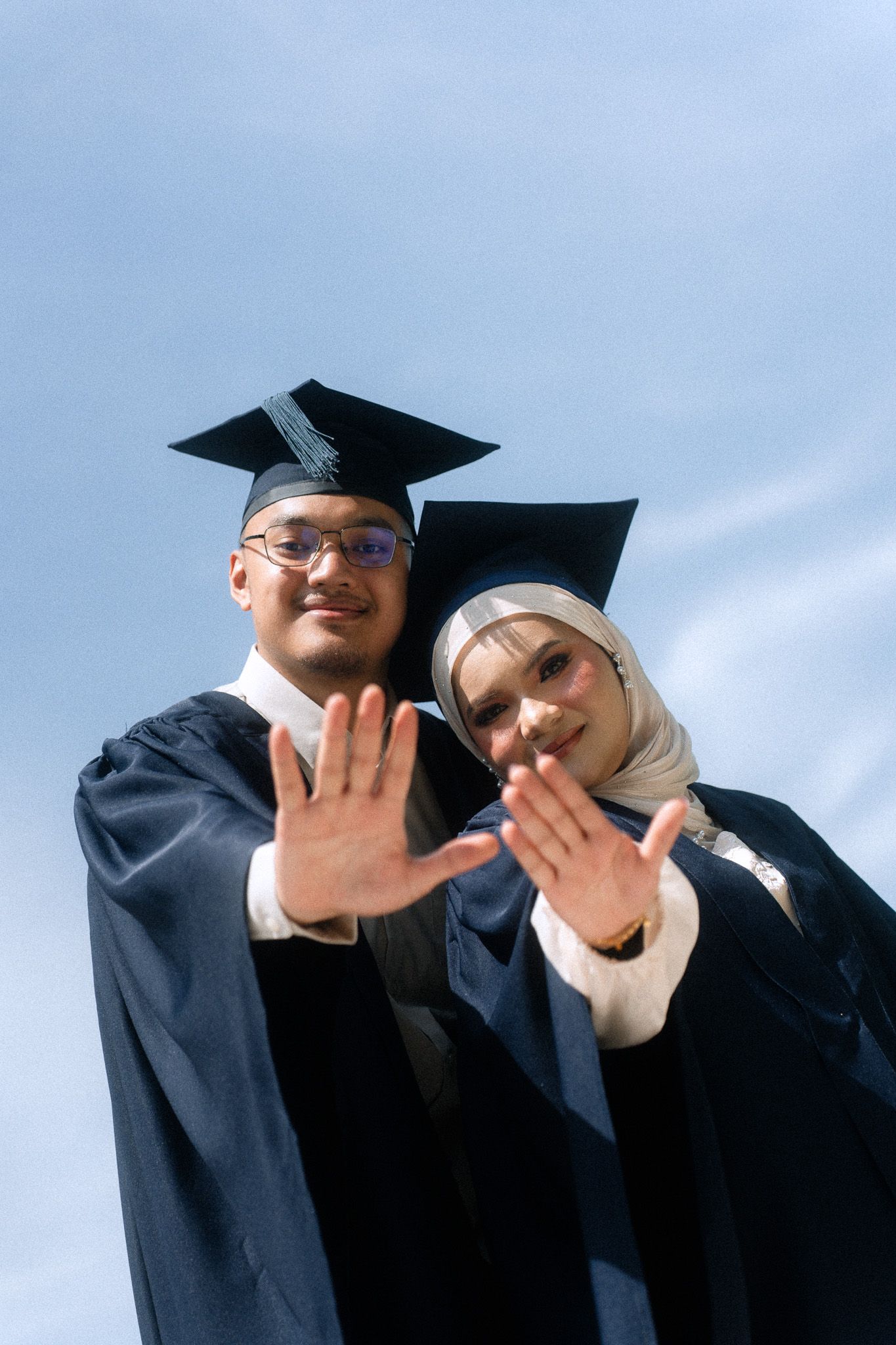 Two graduates in caps and gowns wave at camera against blue sky, one wearing hijab.