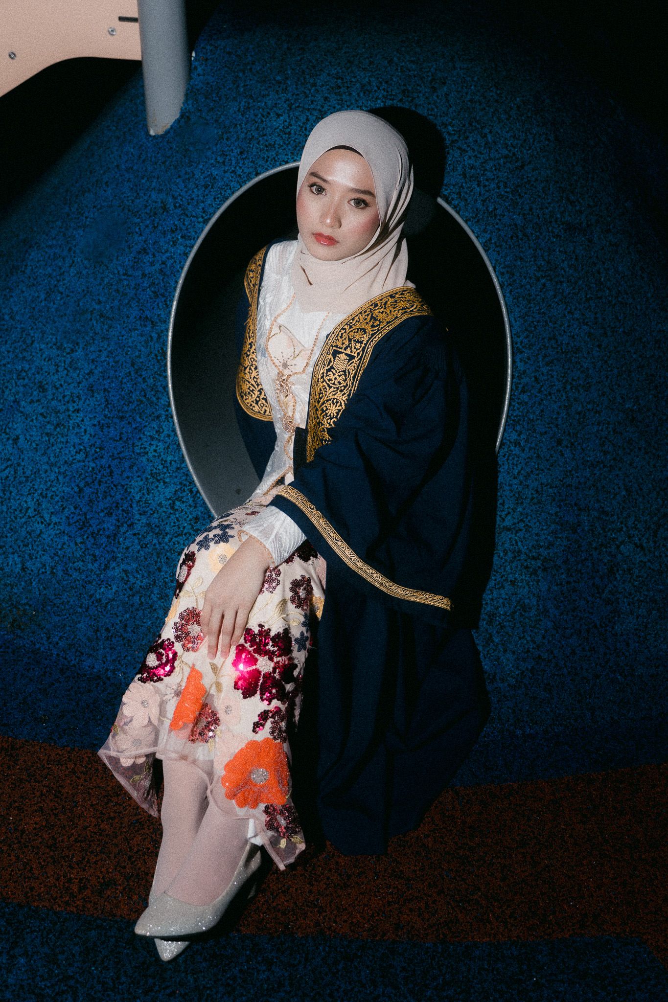 Young Muslim woman in hijab and floral skirt poses in circular mirror frame on blue carpet floor.