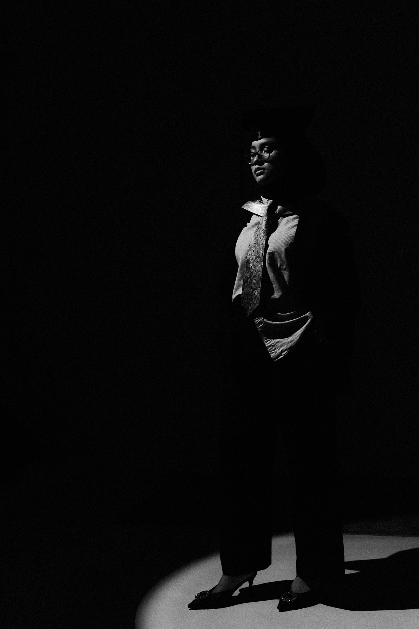 Dramatic black and white full-body portrait of a woman standing in a spotlight against dark background.