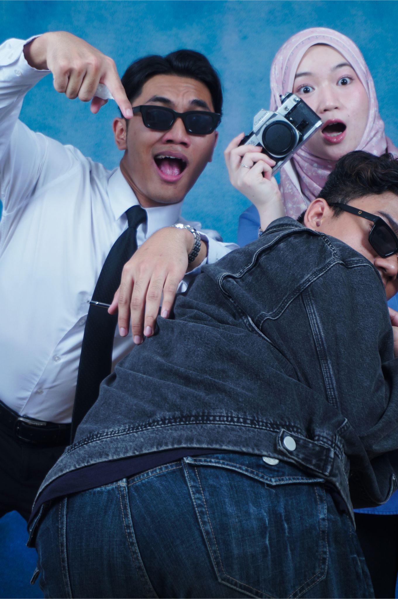 Group of young friends posing playfully with a vintage camera, wearing sunglasses and casual attire against a blue background.