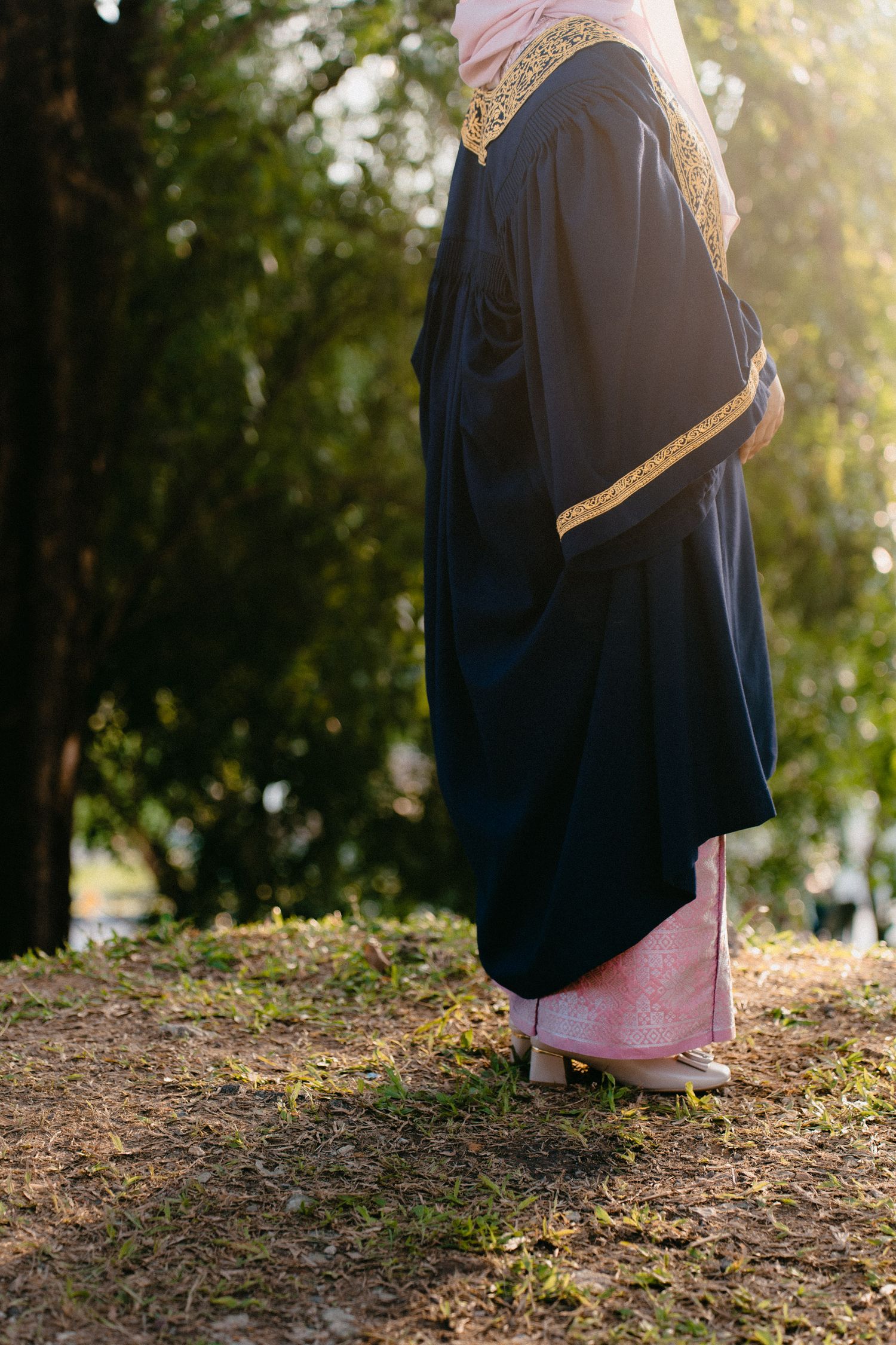 A person wearing a navy blue graduation gown and pink stole stands in dappled sunlight on a grassy hillside.