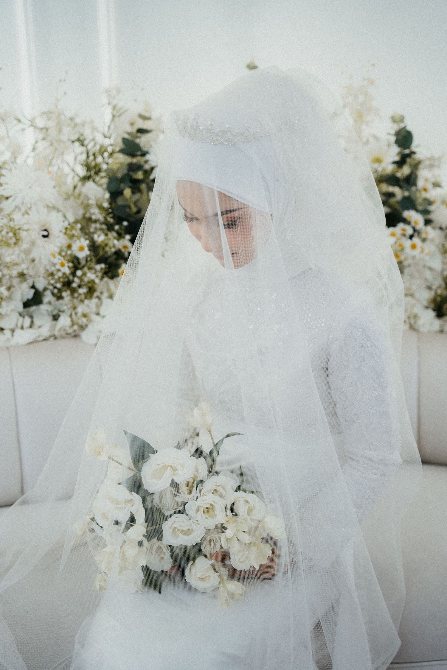 A bride in a white hijab and flowing wedding dress holds an elegant bouquet of white roses against a floral backdrop.