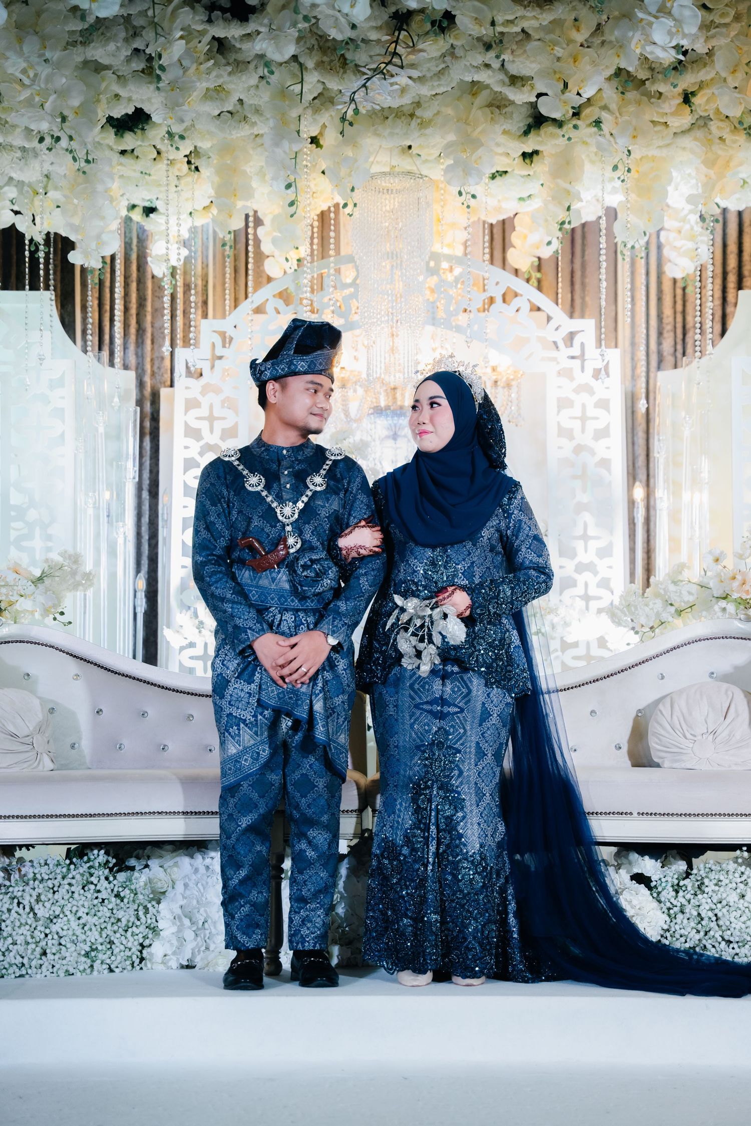 Loving couple in matching navy blue outfits pose together on elegant white sofa in floral-decorated studio setting.
