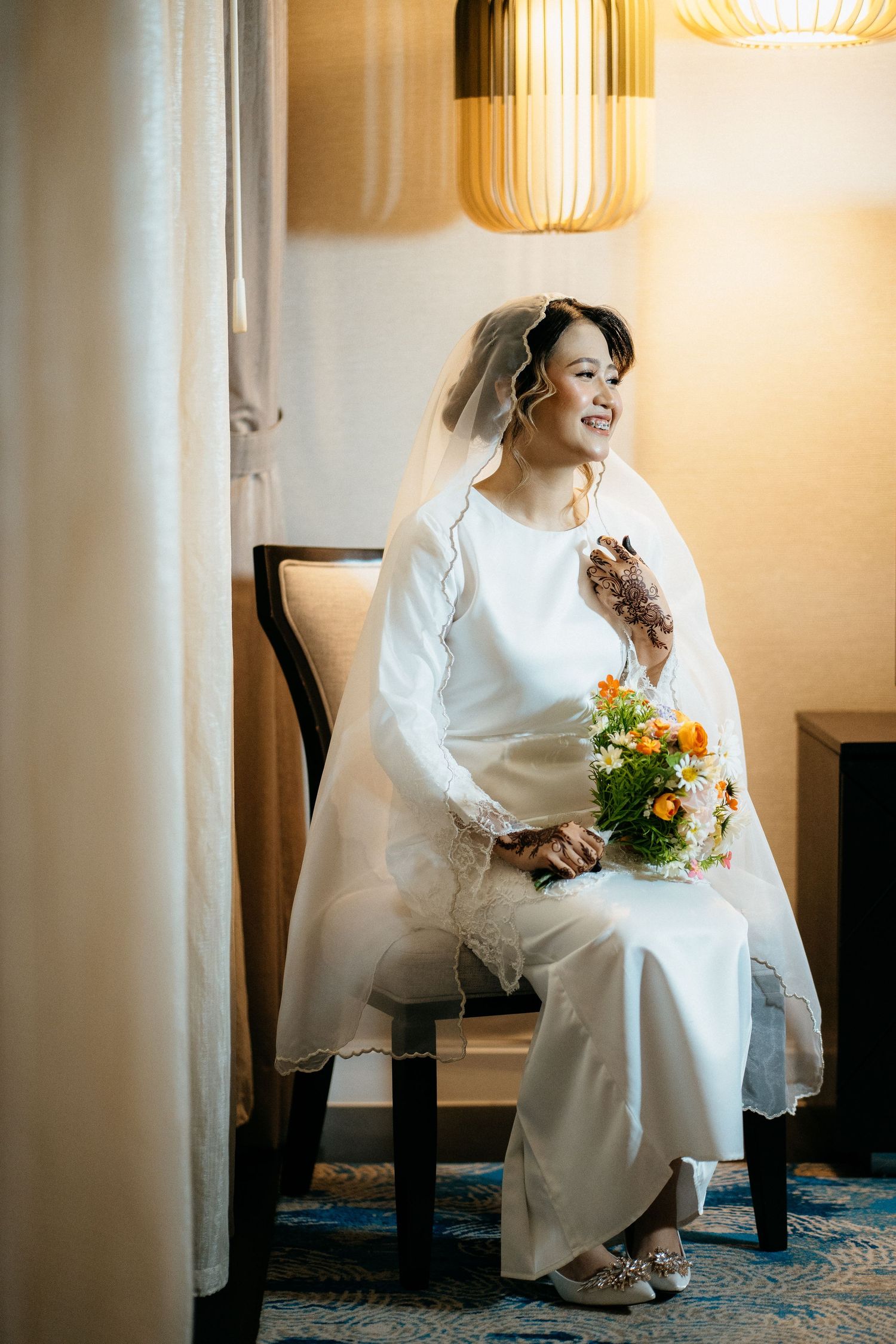 A bride in a white wedding dress and veil sits on a chair under a pendant lamp while holding a bouquet of flowers.